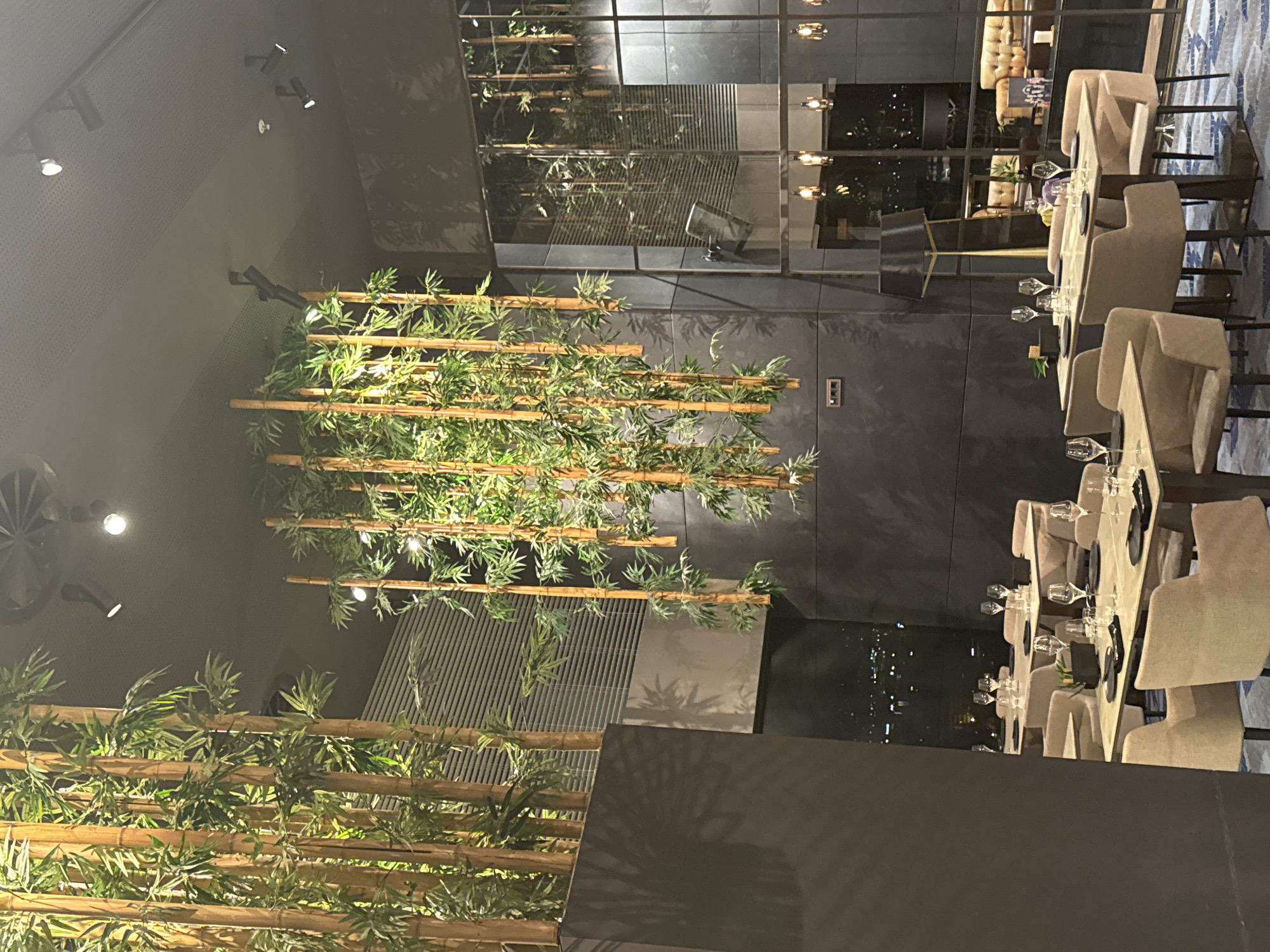 This image shows an indoor dining area in Sofia, Bulgaria, possibly in a restaurant or event space with an Asian-inspired theme. A decorative bamboo installation with live greenery is prominently displayed. Long, rectangular tables are set for dining with glassware, plates, and beige upholstered chairs. Reflections in the room suggest the space may be elevated, perhaps a high-rise, with views of the city lights outside the windows. The setting suggests a planned or staged event. No people are visibly in frame.