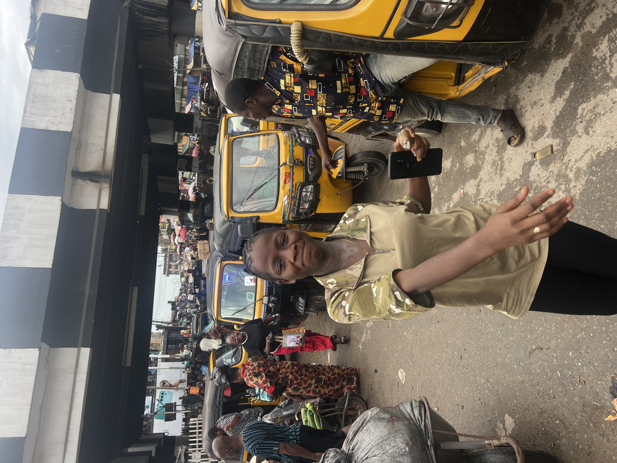 The image depicts a bustling street scene in Nkpor, Nigeria, under an elevated road structure. In the foreground, a young woman with braided hair and a camouflage-patterned shirt is looking at the camera and smiling. She is holding a smartphone in her right hand. Behind her, a man in a colorful, patterned shirt and jeans is leaning out of a yellow auto-rickshaw, seemingly interacting with someone. Several other auto-rickshaws are visible, along with a cart laden with sacks of goods and a trolley with cucumbers. In the background, numerous people are walking and gathered, suggesting a market or busy thoroughfare. The environment is characterized by a paved road, some overhead wires, and buildings lining the street. The overall impression is one of everyday activity in a vibrant urban setting.
