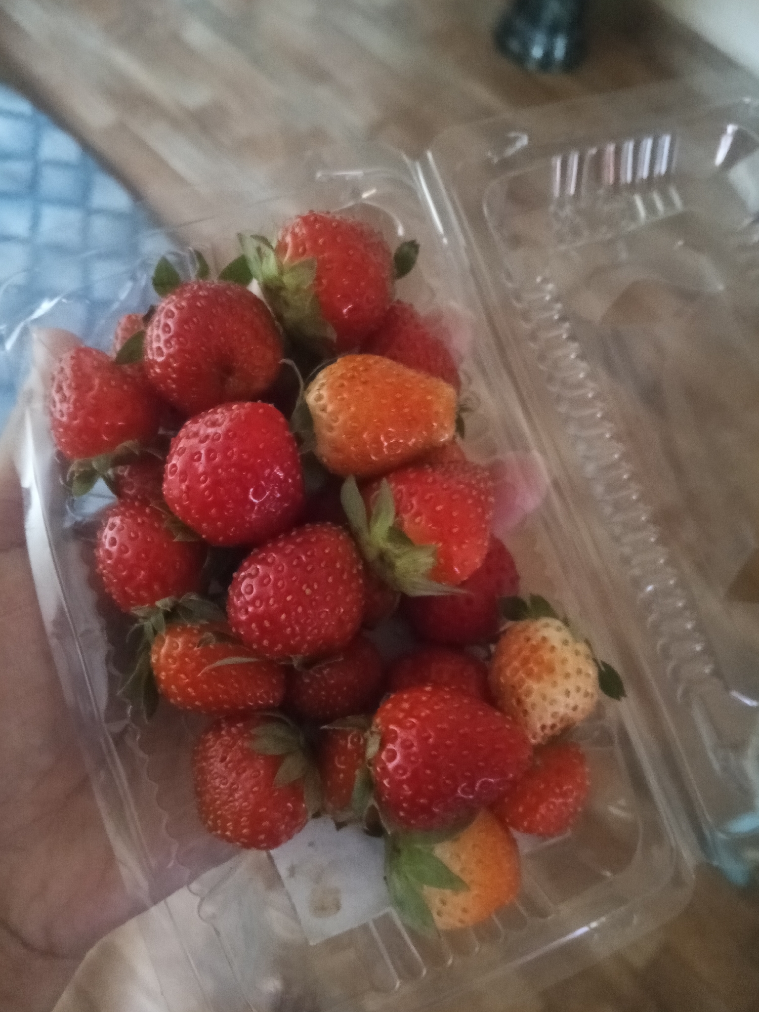 A plastic container of strawberries sits on a wooden surface, possibly a table. The strawberries are varied in ripeness, with some deep red and others a lighter orange hue. The container is partially open, revealing the fruit within. The background is blurred, but appears to be an indoor setting with a hint of blue fabric and natural light. There is no indication of the specific location in Lubuklinggau, Indonesia, or any people present in the image.