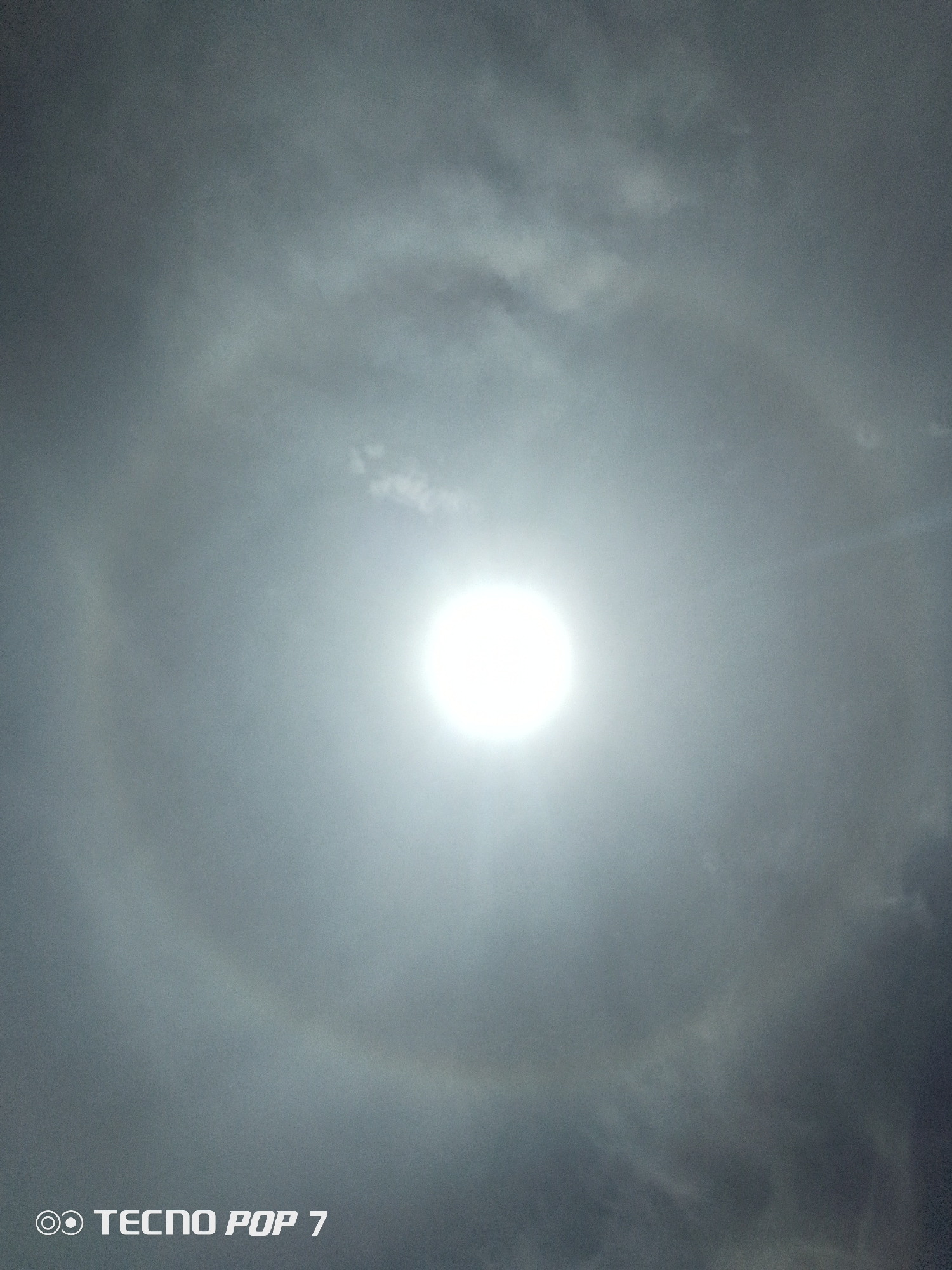 This image presents a bright solar halo observed in an overcast sky over the city of Altagracia, Venezuela. A brilliant, circular light source, consistent with the sun, is centrally positioned in the upper half of the frame, partially obscured by a layer of diffuse, grey clouds. A distinct, wide, circular 22-degree solar halo is visible around the central light source, appearing as a brighter ring against the surrounding grey cloud cover. The sky exhibits varying densities of thin, grey clouds, which diffuse the sunlight. The overall color palette is dominated by shades of grey and white. No individuals, animals, or man-made structures are discernible within the environmental context. A digital watermark, "OO TECNO POP 7," is present in the lower-left corner of the image.