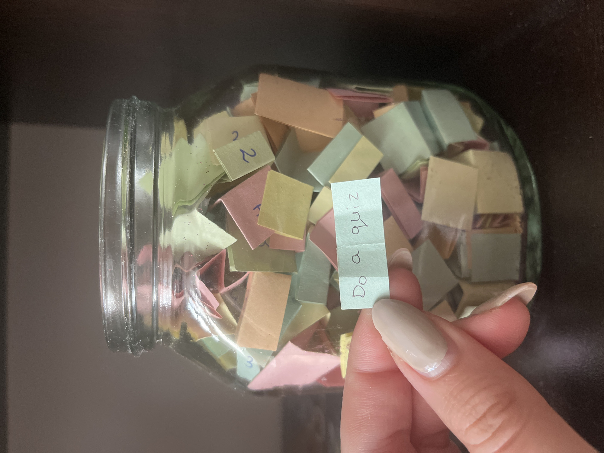 A person holds a small, folded piece of light blue paper with the phrase "Do a quiz" written on it. The paper is being held in front of a glass jar filled with other small, folded pieces of paper of various colors (pink, yellow, green, and white). Some of the papers in the jar have numbers written on them. The jar is sitting on a shelf in front of a dark wooden surface. The person's hand with neatly manicured fingernails is visible in the lower right corner of the image. The scene appears to be indoors. There is no information to suggest this image was taken in the city of Varna, Bulgaria.