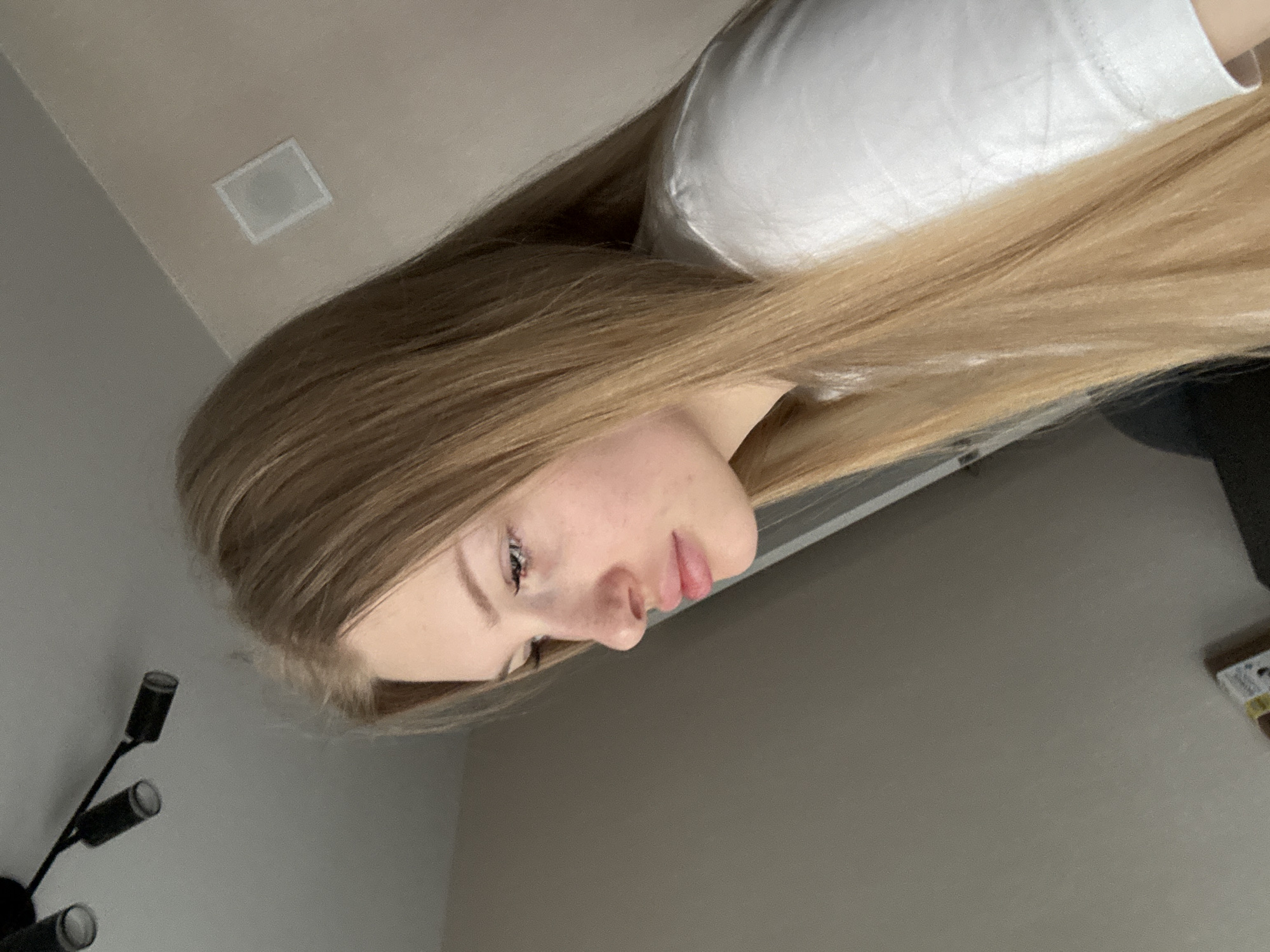 The image shows a young woman with long blonde hair, looking down. She is wearing a white t-shirt. The background appears to be an indoor setting with a textured wall and a ceiling with lighting fixtures. There is also a hint of what looks like a window frame on the right side of the image. The overall scene is indoors and private, with no discernible urban or city elements visible to pinpoint a location within Perm, Russia.