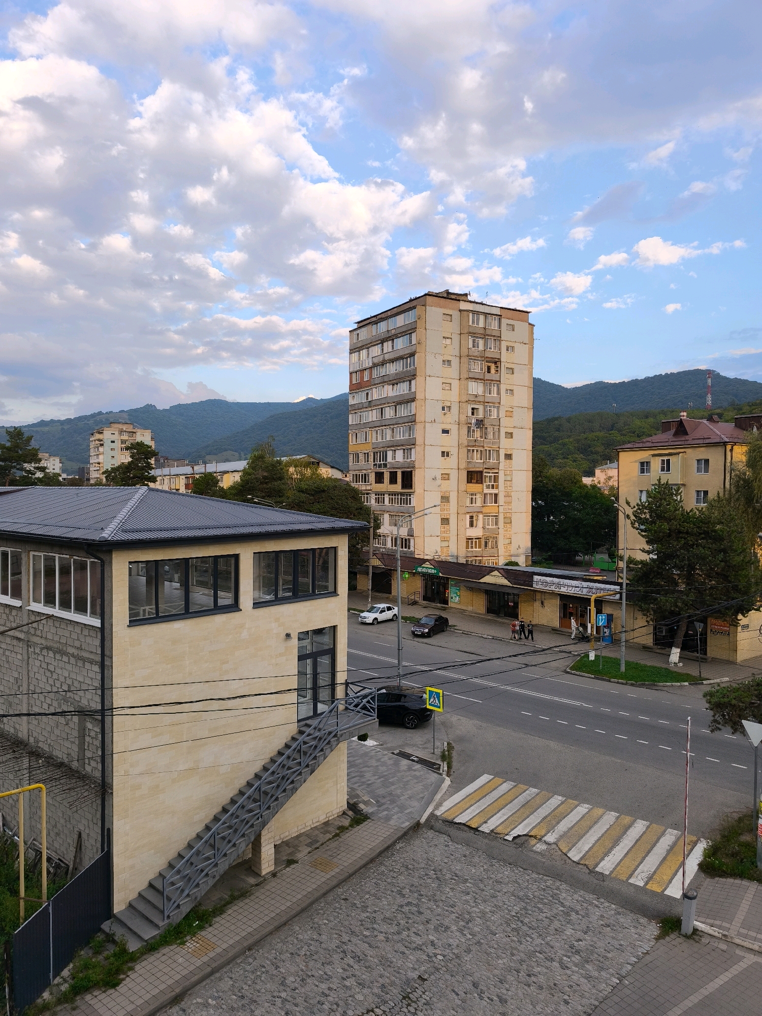 The image shows a view of a street in Karachayevsk, Russia, with a tall apartment building and other structures. The foreground features a modern building with a grey metal roof and large windows, adjacent to an external metal staircase. A cobblestone pathway and a pedestrian crossing with yellow and white stripes are visible on the street. A black car is parked near the crossing, and a yellow yield sign is posted on a pole. Further down the street, a multi-story apartment building with numerous windows dominates the scene, with a yellow-colored building and trees to its right. In the background, lush green hills and mountains under a partly cloudy sky suggest a natural landscape surrounding the urban area. The lighting indicates it is either late afternoon or early morning.