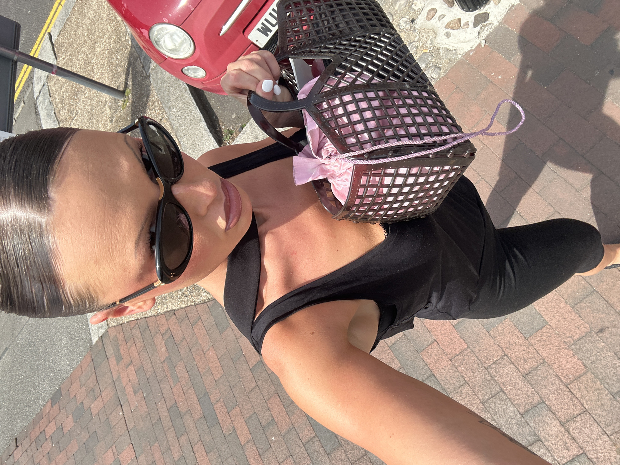 A woman wearing sunglasses and a black dress is holding a black lattice-patterned bag with a pink satin pouch inside. She is standing on a brick-paved sidewalk. In the background, part of a red Fiat 500 car is visible, with the partial license plate "WU". The scene appears to be outdoors in daylight. The location is Tunbridge Wells, United Kingdom.