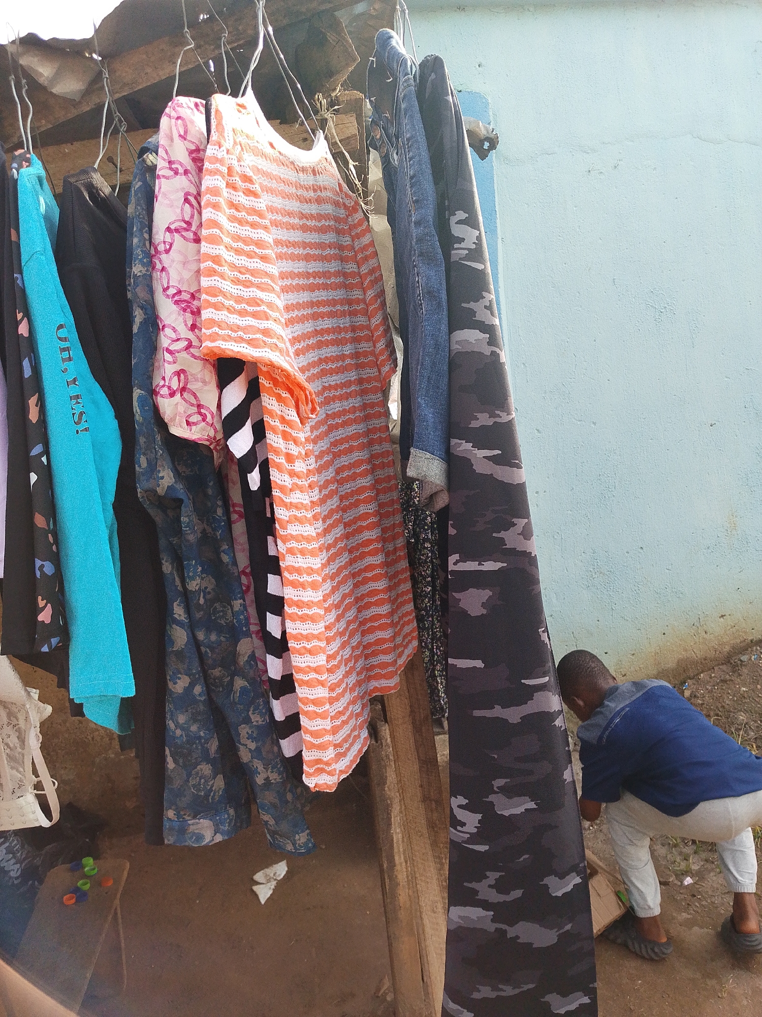 The image displays an outdoor scene, likely in Ohokobe Ndume, Nigeria, centered around a collection of clothing hanging on a makeshift rack. Numerous garments are suspended from simple wire hangers, suggesting either clothes drying or items for sale. Visible clothing includes a bright turquoise long-sleeved top with the text "OH, YES!", a black shirt, a sheer pink and white patterned fabric, a dark blue floral patterned garment, an orange and white striped top, a black and white striped top, blue denim jeans, and a black and grey camouflage patterned fabric. A white lace bra is partially visible on the far left. The rack itself appears to be constructed from rough wooden planks, with a corrugated roof visible above.
In the lower right corner, a person, possibly a child or young man, is partially obscured by the hanging clothes. He is seen from behind, crouching down, wearing a blue short-sleeved shirt, light-colored pants, and dark slides or sandals. The background features a light blue-painted wall. The ground is unpaved dirt. To the left, a small wooden stool or box holds several colorful plastic bottle caps (green and orange). A crumpled white item and a cardboard box are also on the ground near the crouching figure. The natural light indicates it is daytime.