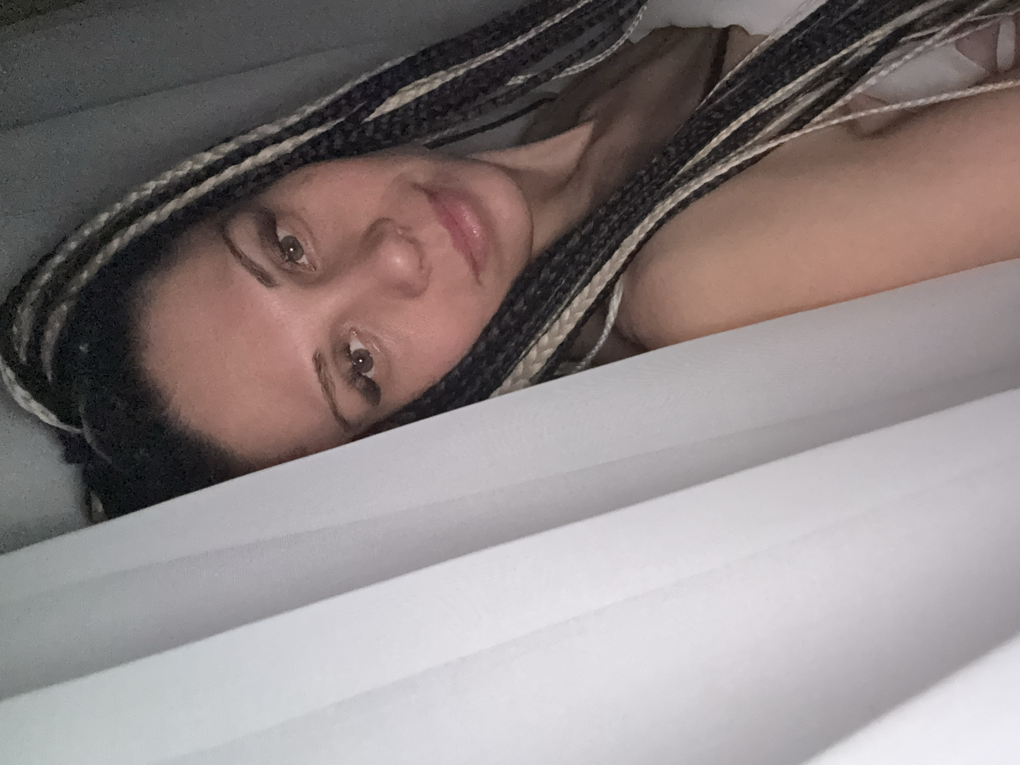The image shows a woman with braided hair lying down. The braids are black and white. She is looking at the camera. She appears to be under white sheets. The location and city are not determinable from the image alone.