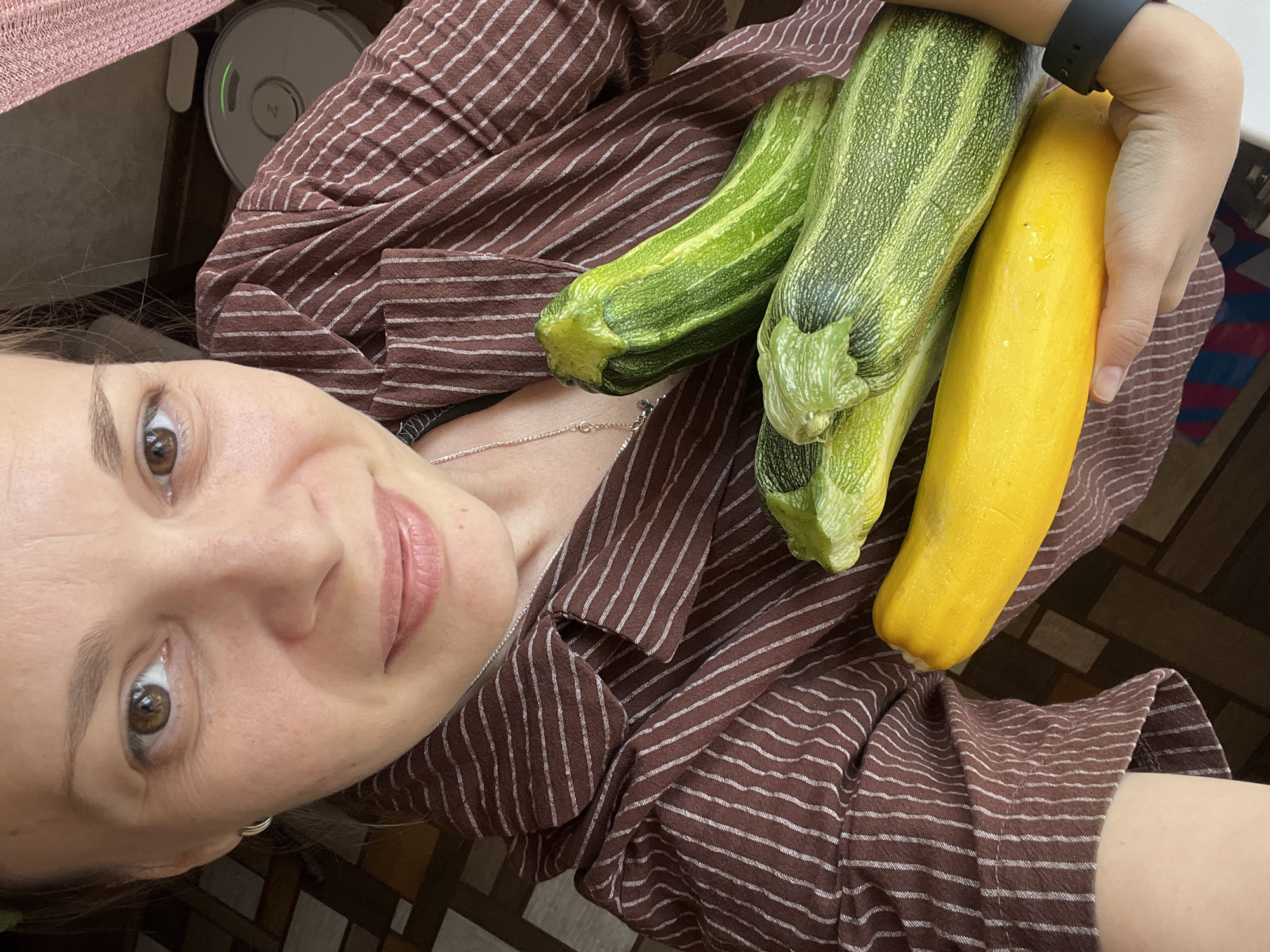 In a close-up, overhead shot, a woman in a brown pinstriped shirt holds a bunch of fresh produce. Her face, partially visible, shows light makeup and a subtle smile. She is holding four zucchini, one bright yellow and three green with lighter stripes. The vegetables are in the foreground, filling the lower half of the frame. In the background, a round, white robot vacuum cleaner with a green light is visible, suggesting an indoor setting. The flooring appears to be tiled with a geometric pattern. The image does not provide any information that allows for determination of the city or country.