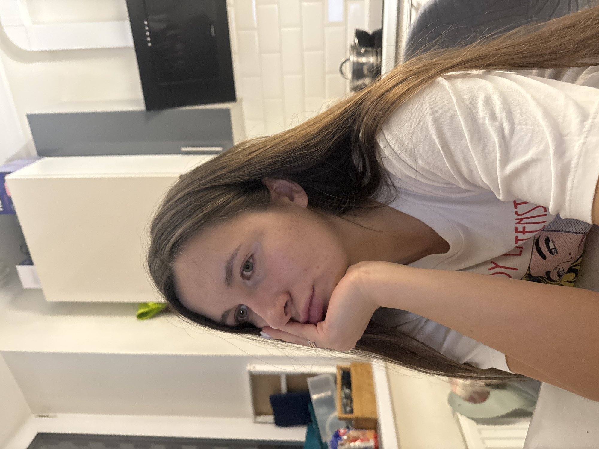 A female individual is depicted indoors, seated at a table within a residential kitchen or dining area in Осоргино, Russia. The subject, possessing long brown hair and fair skin, rests her left hand, adorned with a three-band ring on the ring finger and white nail polish, under her chin. She wears a white short-sleeved t-shirt featuring a partial graphic design. Her gaze is directed towards the left side of the frame. The background reveals a kitchen area with white subway tile backsplash, a black range hood, white and light grey cabinetry, and a metal pot with a lid on a stovetop. To the left, a window or door frame, a white radiator, and a shelving unit containing various items including a blue container and a wooden box are visible.