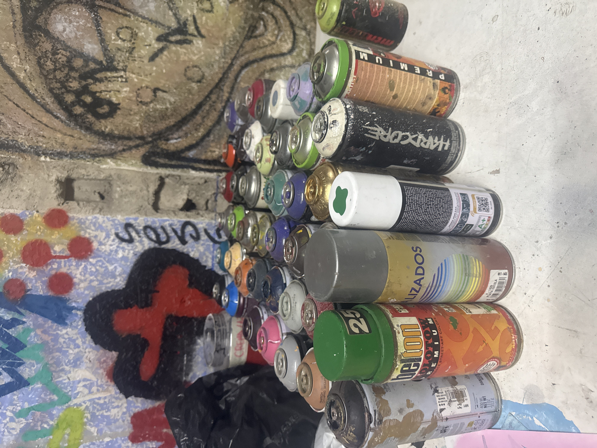 The image shows a collection of spray paint cans on a table, with graffiti art on the wall behind them. The cans are of various brands and colors, and some are covered in paint splatters. The graffiti on the wall is abstract and colorful, with a large black and red shape resembling a deformed heart. The scene appears to be in an urban environment, possibly an art studio or an outdoor graffiti spot. There are no people or animals visible in the image. The lighting is dim and appears to be artificial, casting shadows on the table and cans. The overall impression is one of a creative space dedicated to graffiti art. There is no information to indicate the location is in Madrid, Spain.