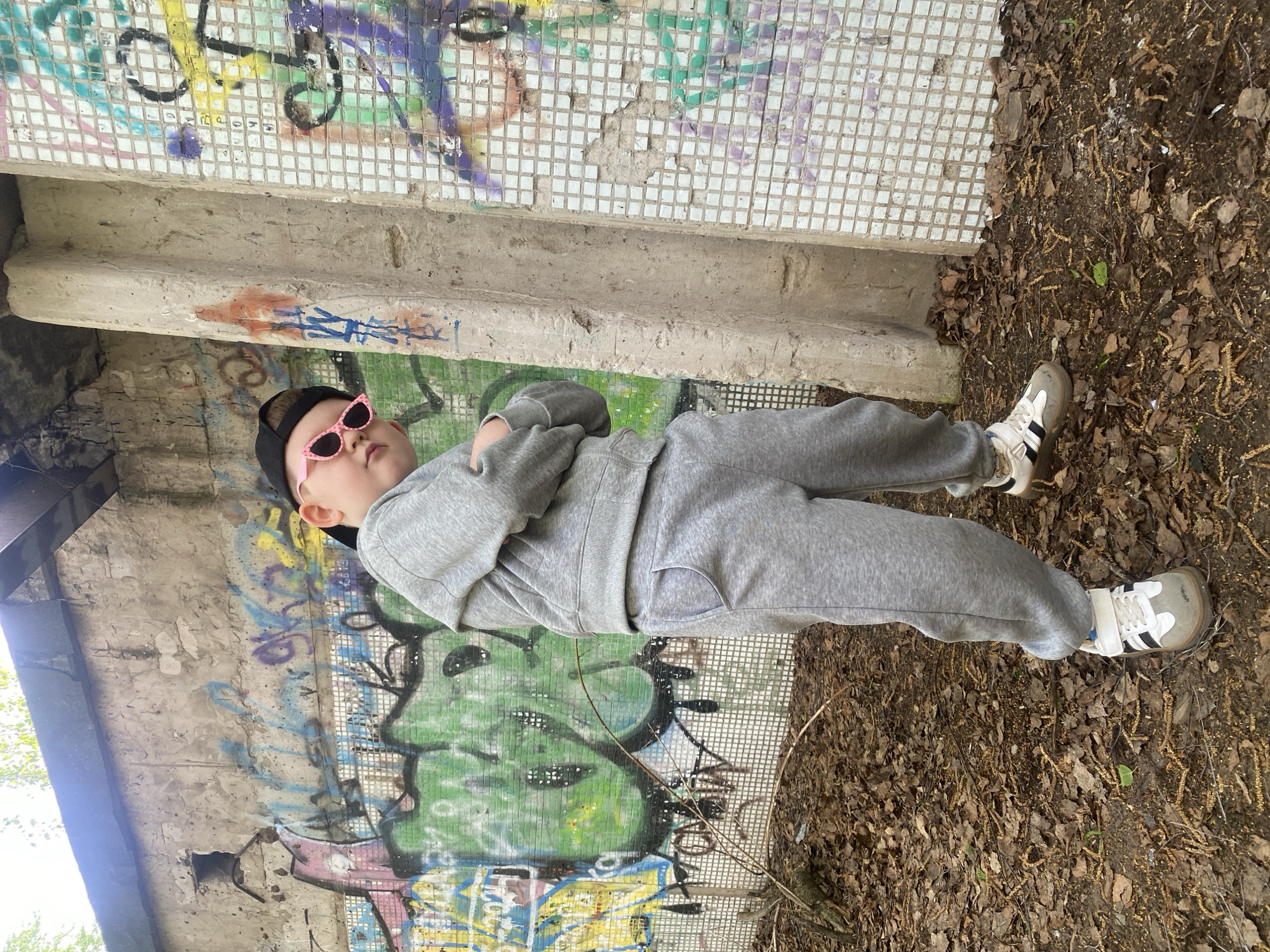 A young person wearing a gray tracksuit, a black cap, and pink sunglasses is leaning against a graffiti-covered concrete structure in a wooded area. The ground is covered in fallen leaves and debris. The graffiti on the concrete includes various colors and shapes. The location is the city of Chaikovsky, Russia.