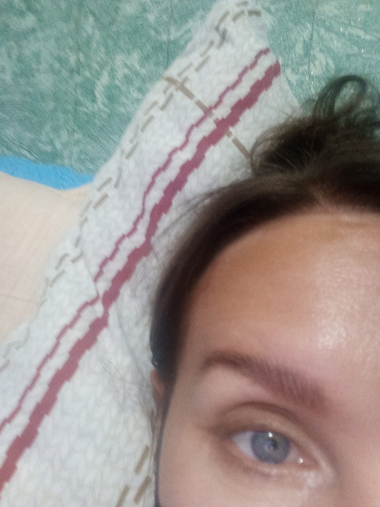 The image shows a close-up of a person's eye and forehead, with a pillow and a textured surface in the background. The pillow has a pattern of pink and beige stripes. The textured surface appears to be a wall or fabric with a greenish hue. The person's eye is light blue, and their eyebrow is dark and well-defined. The lighting is soft, and the image has a slightly grainy texture. There is no information about the location in the city of Novopokrovka, Russia, or any specific actions or interactions taking place.