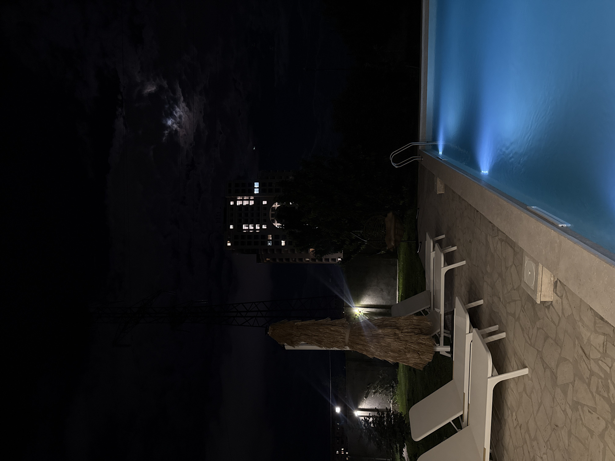 The image depicts a nighttime scene in Yerevan, Armenia, near a swimming pool. The pool is illuminated by blue lights. Adjacent to the pool are several white lounge chairs. In the background, a building with lit windows is visible, along with a lattice structure, possibly a utility tower. The sky is dark with some visible clouds and a faint moon. The scene is lit by artificial lights, including those illuminating the pool and nearby foliage. The ground around the pool is paved with stone slabs.