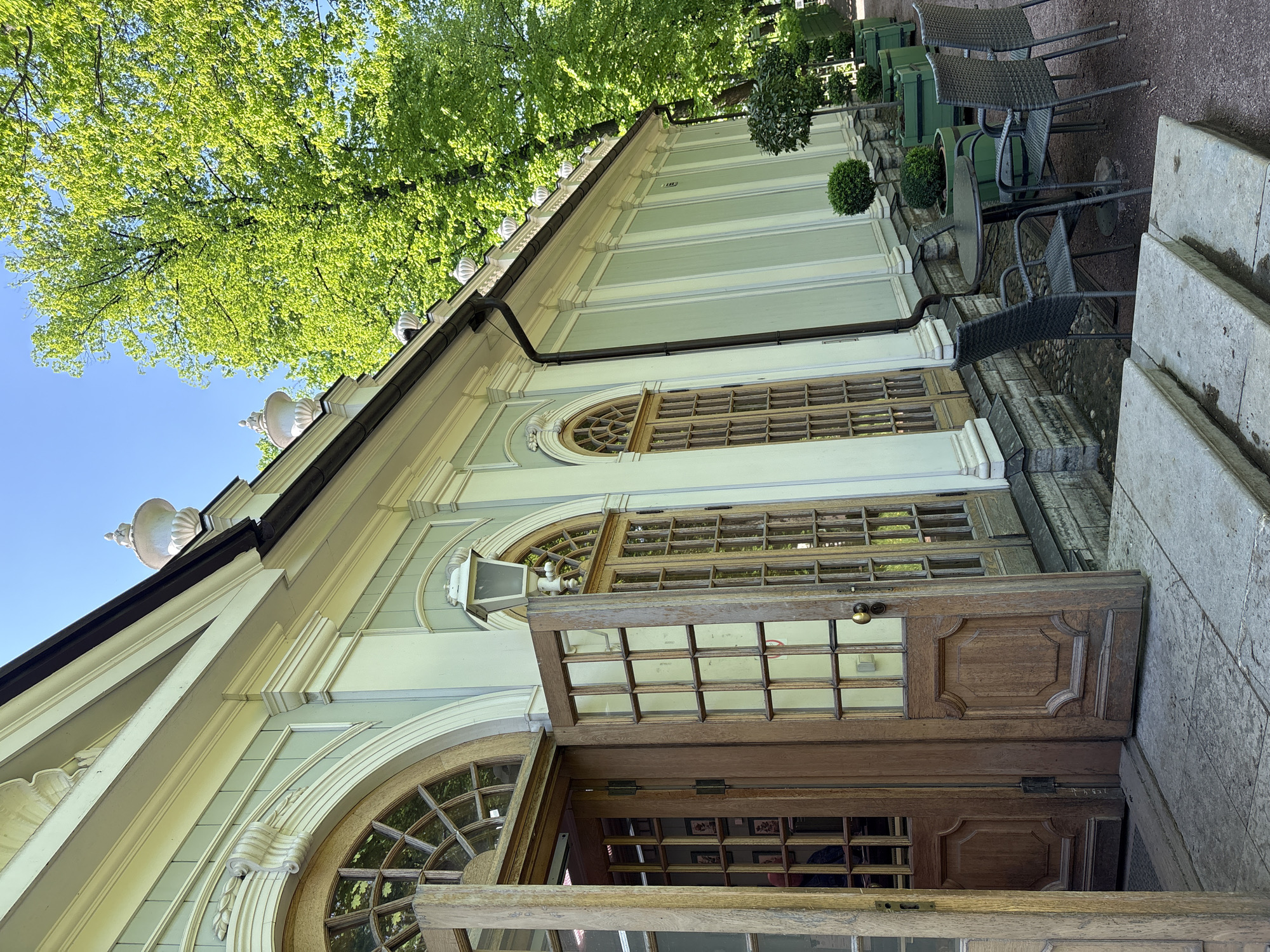 The image shows an exterior view of a building, likely a pavilion or a cafe, situated in Saint Petersburg, Russia, potentially within a park. The building is characterized by its pale green façade with white accents and detailed moldings. Large arched windows with wooden frames are visible, and the doors are open, offering a glimpse inside. A few outdoor tables with chairs are arranged along the side of the building, suggesting a dining area. Lush green trees provide shade overhead. The building's architectural style indicates a historical context, typical of Saint Petersburg's aesthetic.