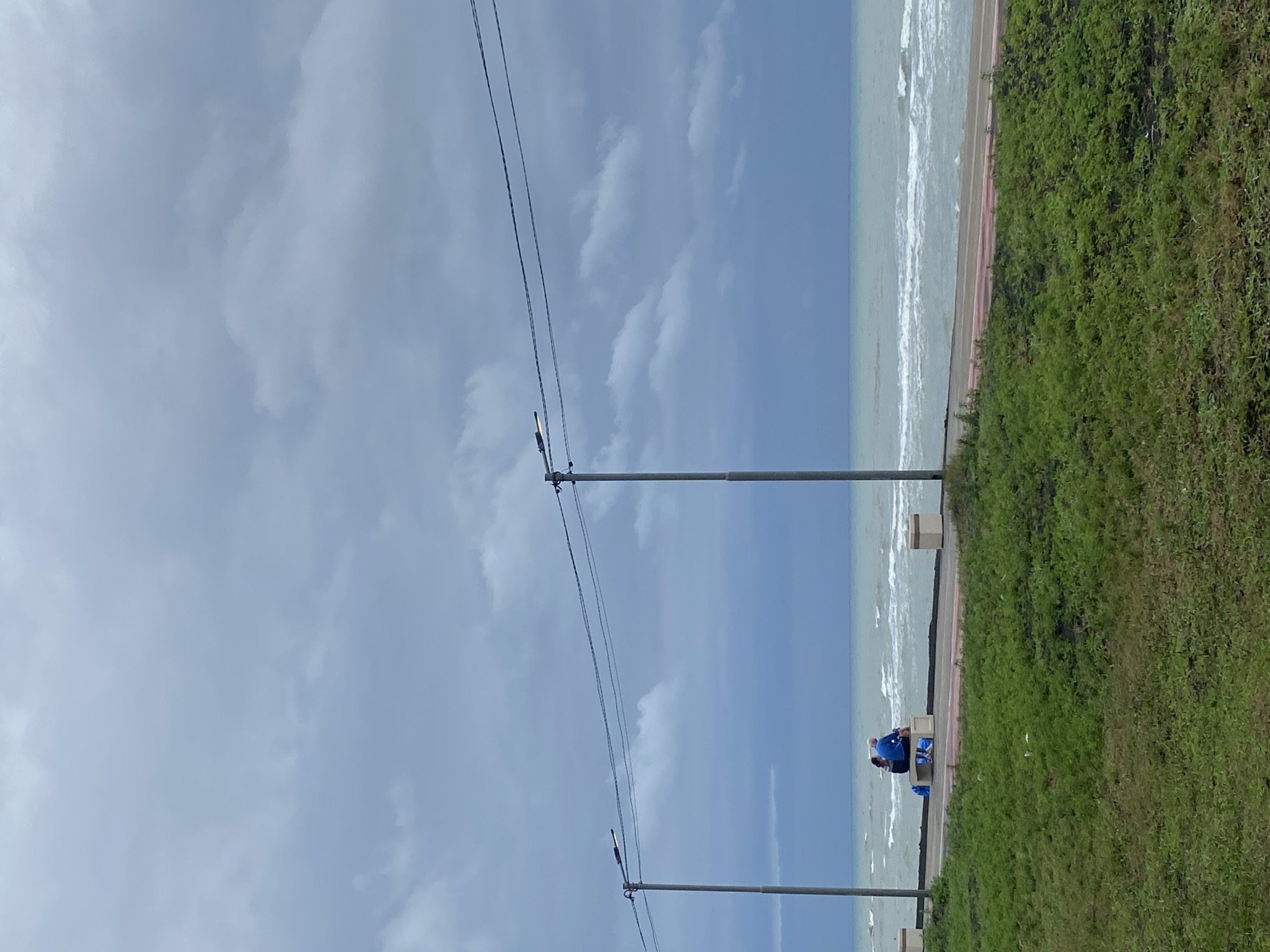 A slightly overcast day at the Ureki, Georgia beach front. Two people, a man and a woman, sit on a bench facing the sea. The man is wearing a blue jacket and the woman is wearing dark clothing. The sea is choppy with white-capped waves rolling onto the shore. A paved walkway with red and white stripes runs along the beach. Two lampposts stand tall with wires connecting them overhead. Green grass covers the foreground of the image.