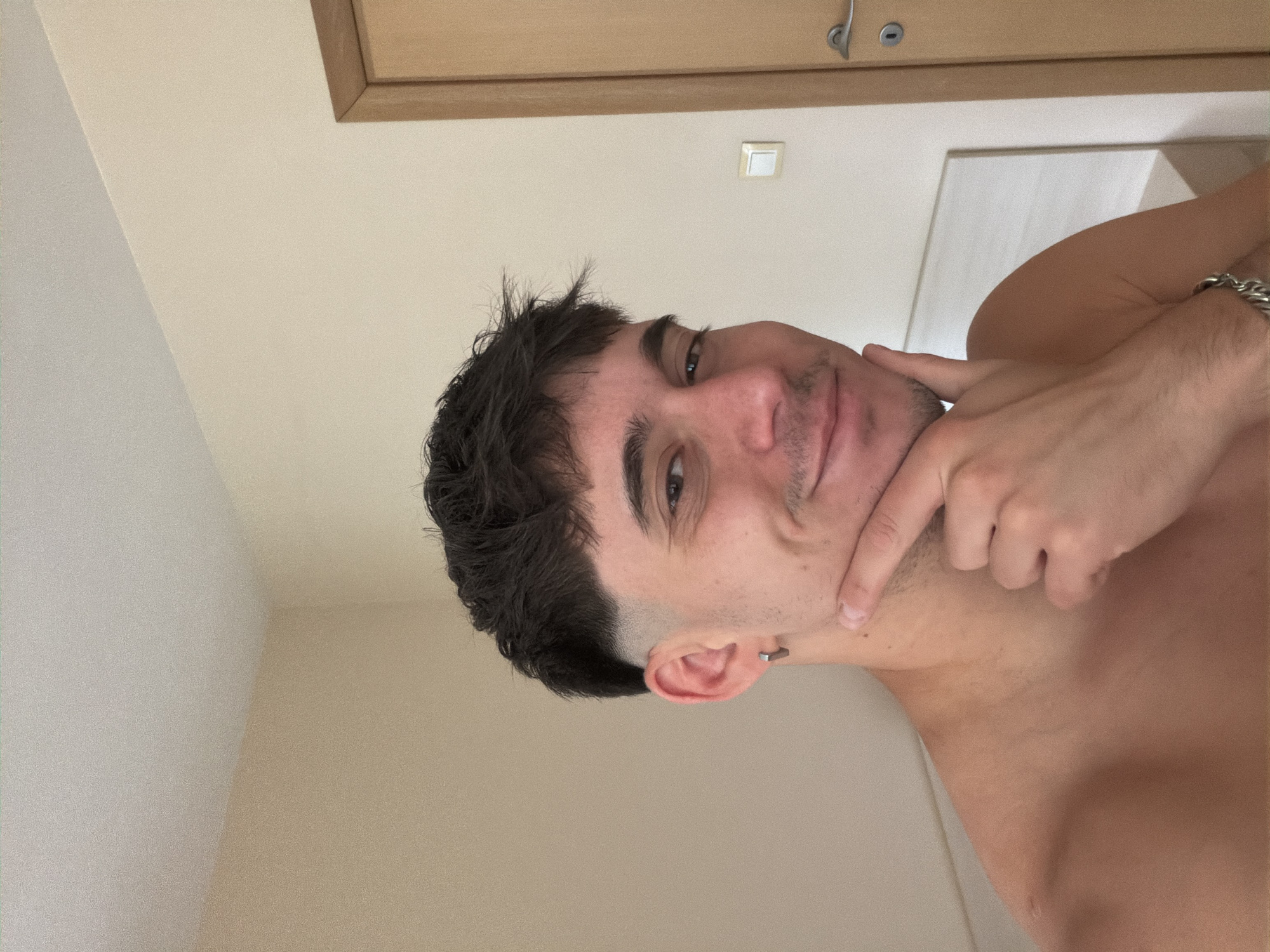 A young man, likely in his early twenties, is featured in a close-up, chest-up portrait, oriented sideways in the original image. He has short, dark, slightly messy hair, dark eyes, and light stubble on his face. He is shirtless, revealing his bare chest and shoulders, and wears a silver chain bracelet on his right wrist. A small, silver hoop earring is visible in his left ear.
He looks directly at the camera with a calm, subtle smile and a slightly contemplative expression. His right hand is raised, with his index finger touching his chin or jawline.
The background consists of light-colored, likely off-white or beige walls, suggesting an indoor setting. In the upper right corner, a wooden door frame or cabinet edge is visible, along with a white rectangular light switch or outlet on the wall. The lighting is soft and even, consistent with an indoor daytime setting. There is no visible text in the image. While the image itself does not contain specific visual cues to determine a city, the provided context places this scene in Blagoevgrad, Bulgaria.