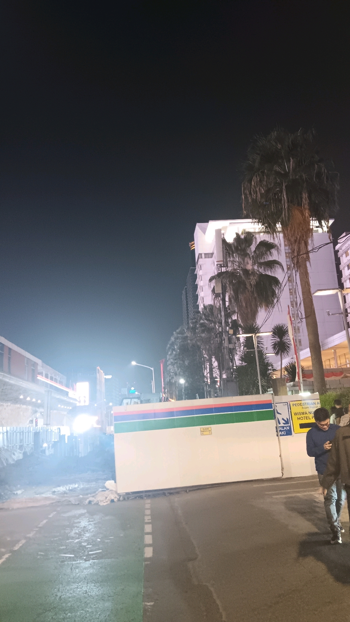 The image depicts a street scene at night in Central Jakarta, Indonesia. In the foreground, a wet asphalt road with white dashed lines marking lanes is visible. To the right, two individuals are walking away from the camera. One person, wearing a blue sweater and jeans, is looking at their phone. Behind them, a white barrier with green and blue stripes runs horizontally across the scene. A signpost is attached to the barrier, with text that is partially legible. In the mid-ground, a tall palm tree stands prominently to the right, with a modern white building featuring multiple windows and balconies behind it. Several other buildings and palm trees are visible in the background, contributing to an urban landscape. The sky is dark, indicating nighttime, with some faint light emanating from streetlights and building illumination. To the left of the frame, a structure that appears to be a railway or elevated walkway is visible, with bright lights suggesting ongoing activity or construction. The overall scene conveys a sense of urban nighttime ambiance with elements of construction and pedestrian movement.