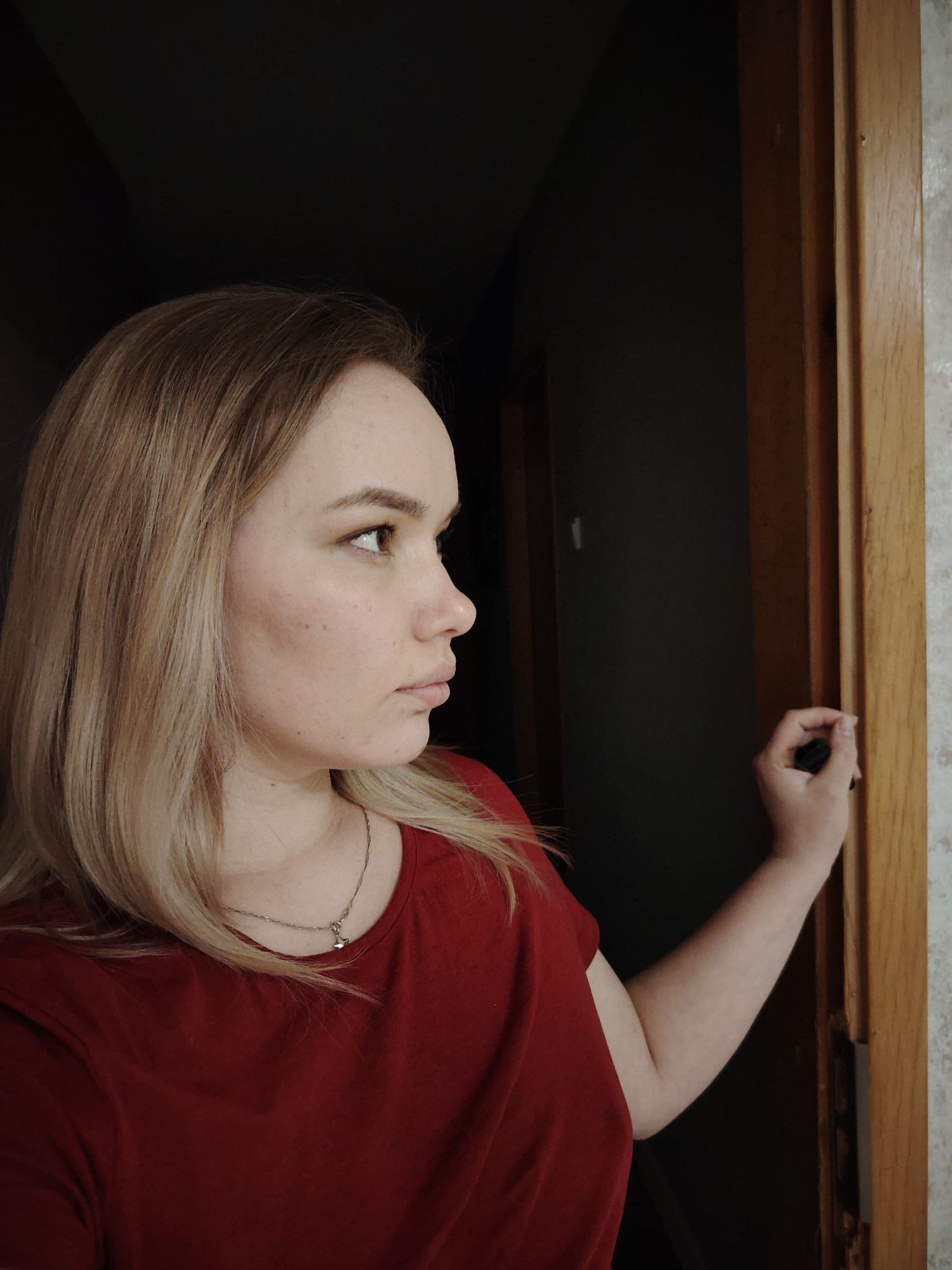 An image taken in Марковский, Russia, depicts a young adult female captured in a head and upper torso profile shot, facing right. She possesses a fair complexion, light brown to blonde shoulder-length hair, and appears to be wearing a plain red short-sleeved t-shirt. A thin silver-colored chain necklace with a small pendant is visible around her neck. Her right arm is bent, and her right hand is grasping a vertical light-brown wooden door frame located on the right side of the image. Her gaze is directed towards the right. The background to her left is deep black, indicating a dark interior, while a lighter, textured surface is visible beyond the wooden door frame on the far right. The lighting emphasizes her left side, casting her right side and the interior space behind her into shadow.