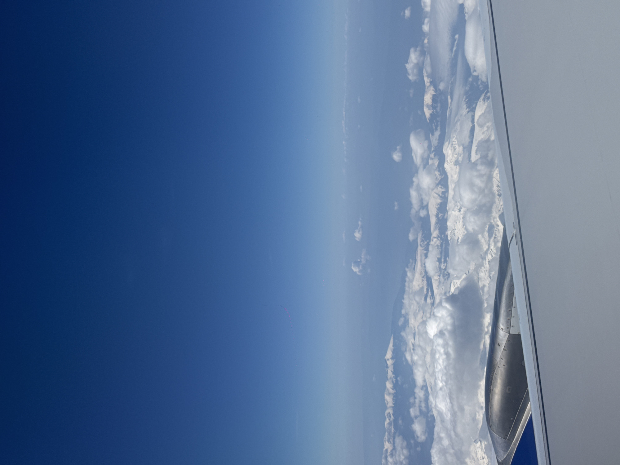 This media file presents an aerial view from an airplane window, showcasing a vast expanse of deep blue sky, an airplane wing, a jet engine cowling, and a landscape of snow-capped mountains partially covered by clouds.
The left side of the frame is dominated by a clear, vibrant blue sky, transitioning from a darker hue at the top to a lighter blue towards the horizon. In this upper sky, a faint, curved trail of small reddish-pink dots can be observed. The right side of the image features the leading edge of a white airplane wing, with a dark, metallic engine cowling visible below it. Beneath the wing and engine, a layer of white, fluffy clouds stretches across the scene, interspersed with rugged, snow-covered mountains.
The image captures the activity of air travel during bright daylight hours. The weather appears clear above the aircraft, with a distinct cloud layer obscuring parts of the mountainous terrain below. Given the provided context of "Maritsa, Bulgaria," the aircraft is likely flying over mountainous regions of Bulgaria, such as the Balkan Mountains or the Rhodopes, which are known for their snow-capped peaks. No discernible text is visible in the image.