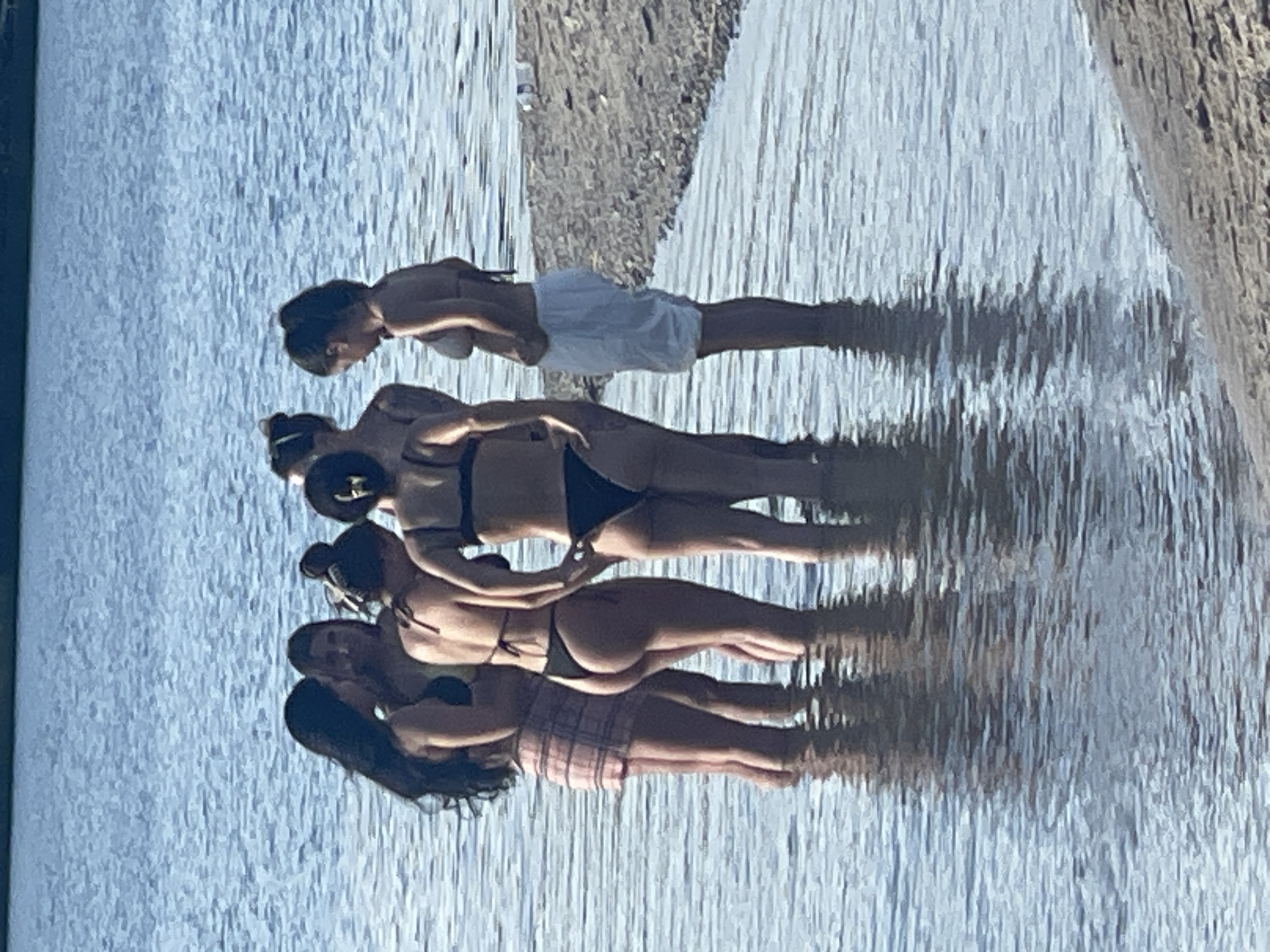 In the city of Unknown, Unknown, six young women are standing in shallow water near a sandy shoreline. They are all wearing swimsuits. From left to right, the first woman has long dark hair and is wearing a black bikini top and a plaid skirt. The second woman has dark hair and is wearing a yellow bikini top. The third woman has dark hair pulled back and is wearing a black bikini top and bottom. The fourth woman has dark hair pulled back and is wearing a black bikini top and bottom. The fifth woman has dark hair pulled back and is wearing a tan bikini top and white shorts. The sixth woman has dark hair and is wearing a black bikini top and bottom. The water is calm and reflects the blue sky. A sandy beach is visible in the background.