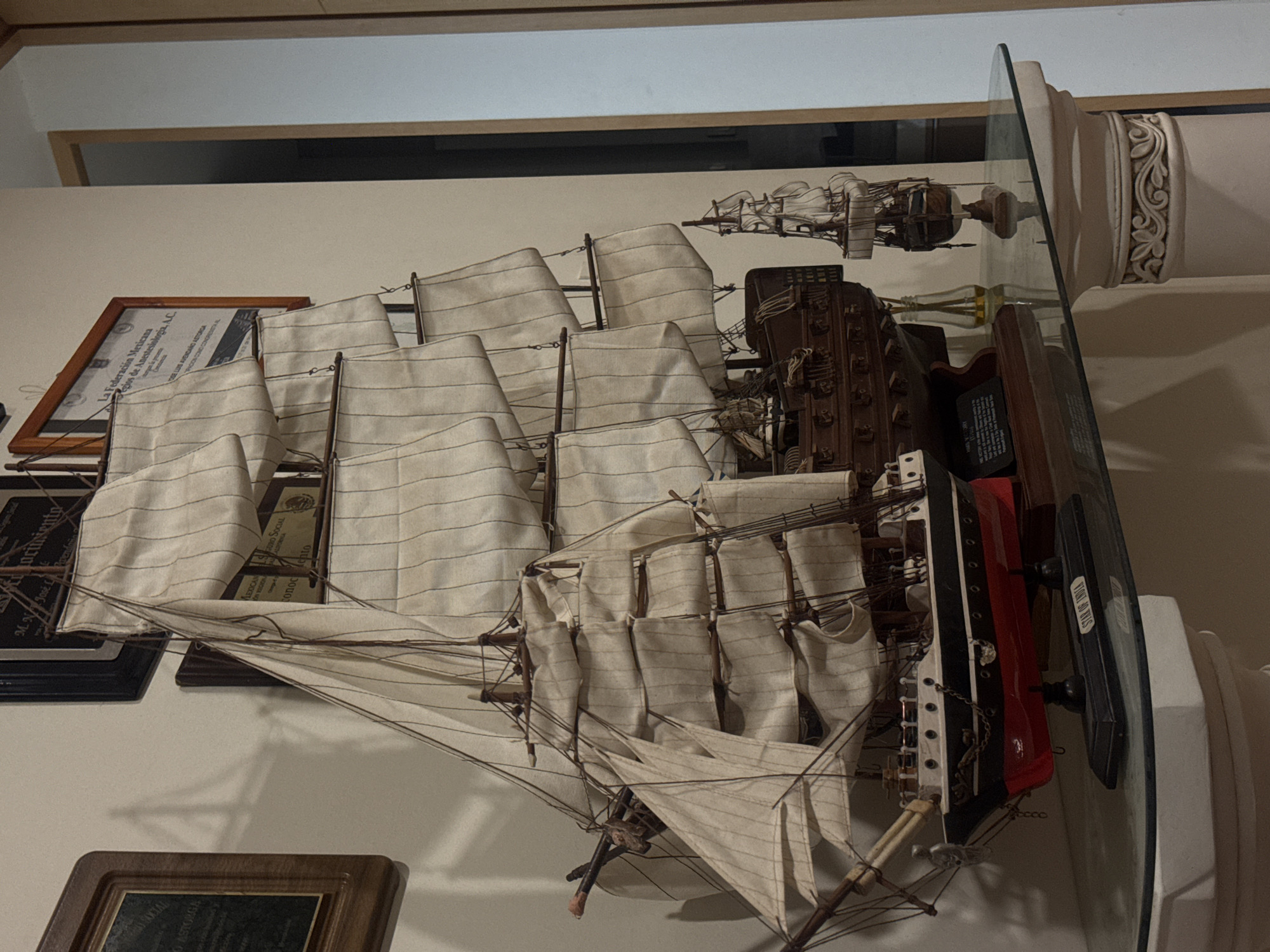 An indoor scene, depicted in a rotated image, showcases a collection of display items, primarily a large, detailed model ship. The main model ship, oriented horizontally (if the image were rotated 90 degrees clockwise), dominates the center. It features multiple off-white sails, intricate brown rigging, and a hull painted black above a red waterline. A small sticker on its base clearly identifies it as "STAR OF INDIA".
The ship rests on a dark rectangular base, which in turn sits on a clear glass shelf. This shelf is supported by an ornate white column-like structure with carved floral patterns, positioned on the right side of the frame. Above and to the right of the main model, on a higher level or shelf, another smaller model ship with sails is visible.
On the light-colored wall behind the display, several framed documents and plaques are mounted. Visible text on these documents includes "La Federación Mexicana de Colegios de Anestesiología, A.C.", "JOSE LUIS AVENDAÑO ASTORGA", and fragmented words like "ASTORGA", "Cincuentena" (Fiftieth Anniversary), "conocimiento" (knowledge), "LEXICAN", "ON VEGION", "URD SOCIAL", and "CALIFORNIA". These suggest professional achievements or memberships. Another wooden-framed plaque is partially visible at the bottom.
The setting appears to be a display area in a home or office, possibly in Puebla, Mexico, given the provided context. The lighting suggests an indoor environment. There is no overt activity taking place, only the static display of these decorative and commemorative items.