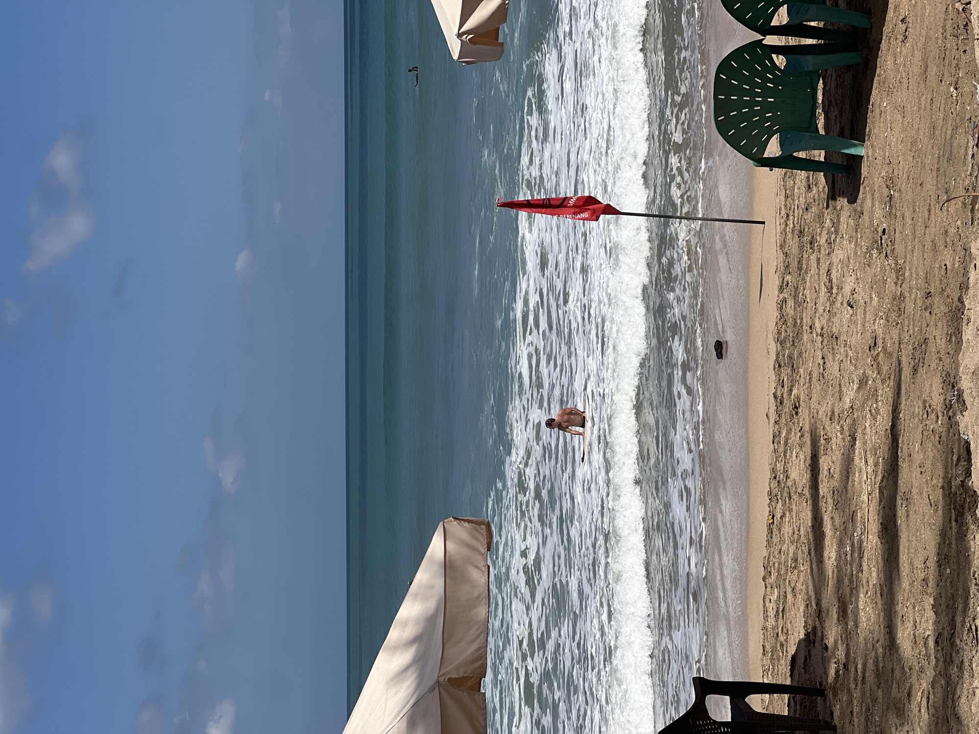 The image depicts a beach scene in Kuta, Indonesia. In the foreground, two empty green plastic chairs are positioned on the sandy beach, casting shadows. To the left, a black plastic chair is partially visible. A red flag on a pole stands near the water's edge. The ocean is visible with waves breaking on the shore. A lone figure is sitting on a surfboard in the shallow water, facing the camera. Further out in the ocean, another individual is on a surfboard. Two large beach umbrellas, one on the left and one on the right, provide shade. The sky is blue with scattered white clouds. The overall scene suggests a relatively calm day at the beach.