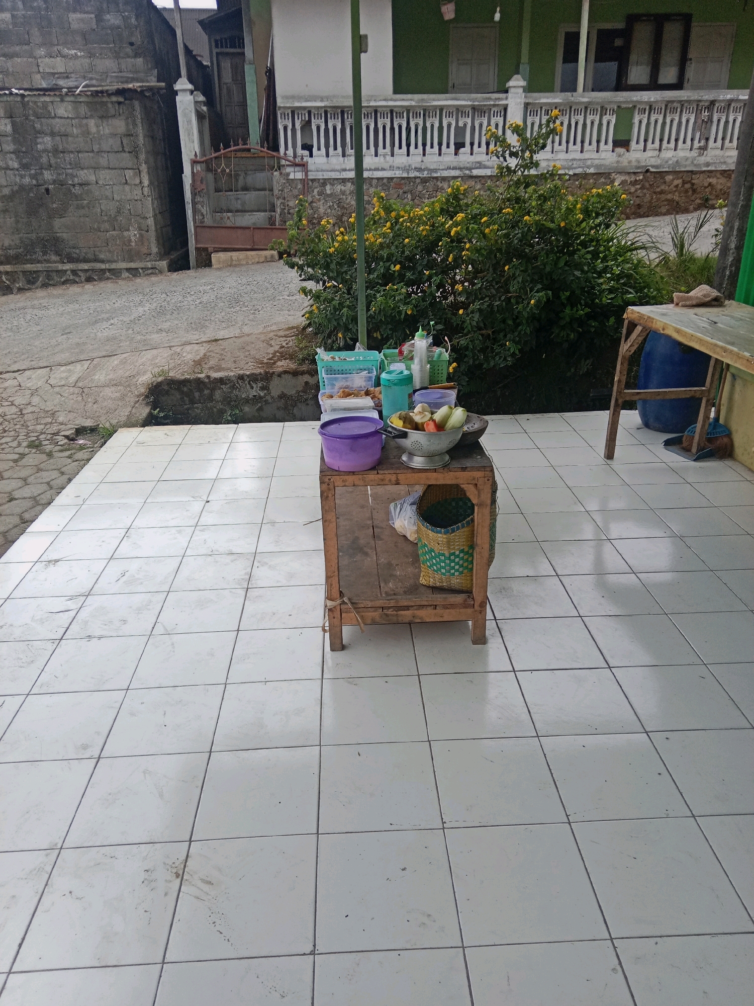 The image depicts an outdoor scene on a tiled patio in Batur, Indonesia. In the foreground, a small, rustic wooden table stands at an angle, displaying a variety of items. On the table, a silver colander holds an assortment of fruits and vegetables, including what appear to be tomatoes, cucumbers, and apples. To the left of the colander is a purple bucket, and to the right, a woven basket with a checkered pattern rests on the table. Below the table, a plastic bag is visible.
Behind the table, a lush green bush with yellow flowers is prominent. Further back, a building with a light green facade and white railing is visible, along with parts of other structures and a weathered brick wall. A dirt road or pathway runs alongside the patio. To the right of the scene, a larger wooden table is partially visible, with a dark blue barrel and a broom leaning against it. The overall impression is a simple, perhaps temporary, roadside stall or an outdoor food preparation area.