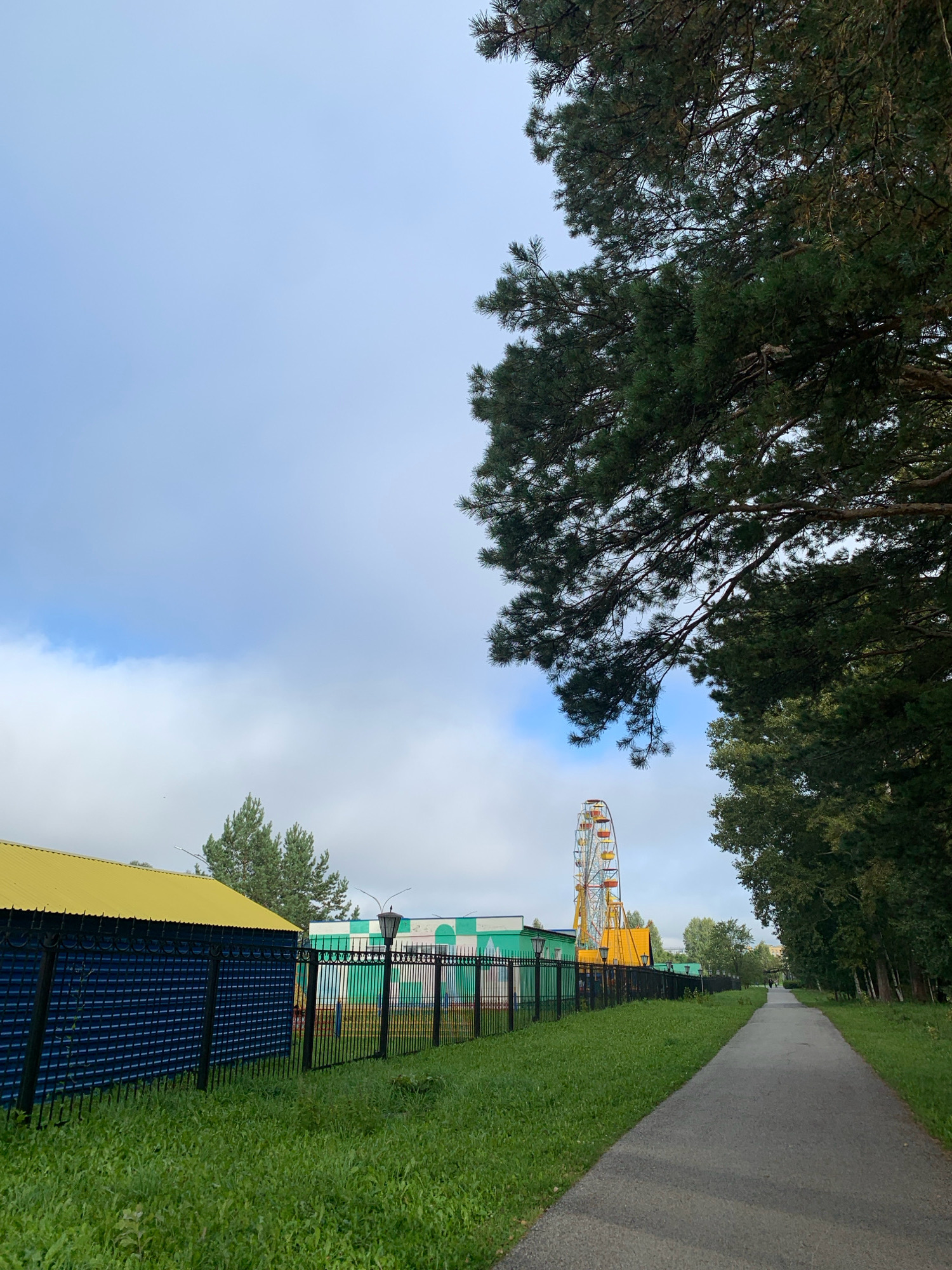 The image shows an outdoor scene in Prokopyevsk, Russia, featuring a park-like area adjacent to an amusement park. In the foreground, a paved pathway runs diagonally from the bottom right towards the center of the image. To the left of the pathway is a stretch of green grass, followed by a black metal fence. Beyond the fence, there are several structures with colorful roofs and walls, including a building with a yellow roof and a blue facade, and other buildings with green and white coloring. A prominent Ferris wheel, painted in yellow, red, and orange, stands in the background, indicating the presence of an amusement park. Tall trees, primarily evergreens and deciduous trees, line the right side of the pathway and also appear behind the park structures. The sky is partly cloudy, with patches of blue sky visible. The overall impression is a daytime scene in an urban or suburban park area that borders an entertainment venue.
