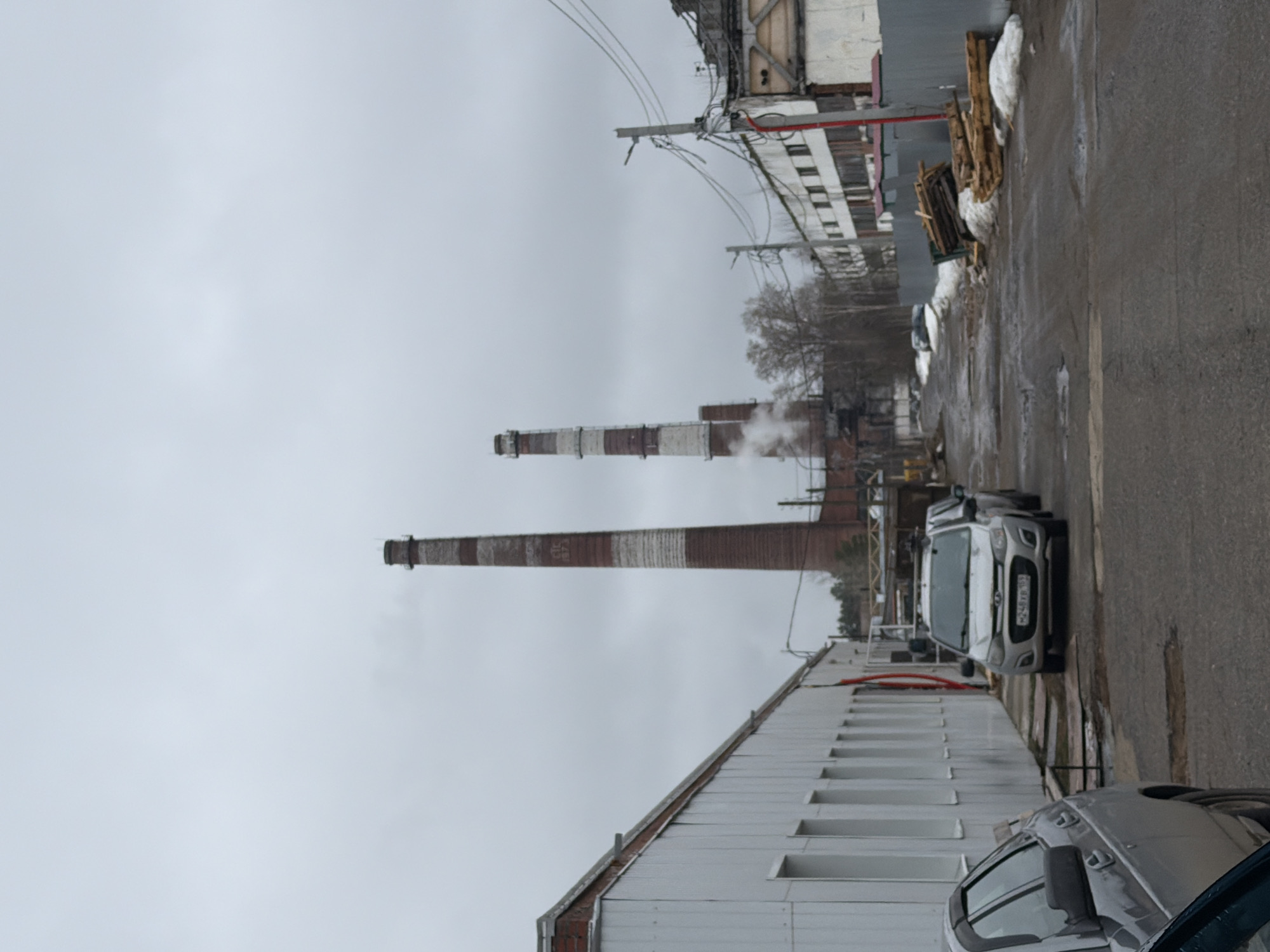 The image depicts a street scene in the city of Chaikovsky, Russia, on an overcast day. Two tall, industrial smokestacks are visible in the background, marked with alternating red and white stripes. One of the smokestacks appears to be emitting smoke or steam. The street is lined with industrial buildings, one of which is white with several windows. A light-colored commercial van is parked on the street near the sidewalk, and the partial view of another vehicle is in the foreground. There is some wooden debris stacked against the side of a building across the street. The road surface appears to be wet and the sky is uniformly gray.