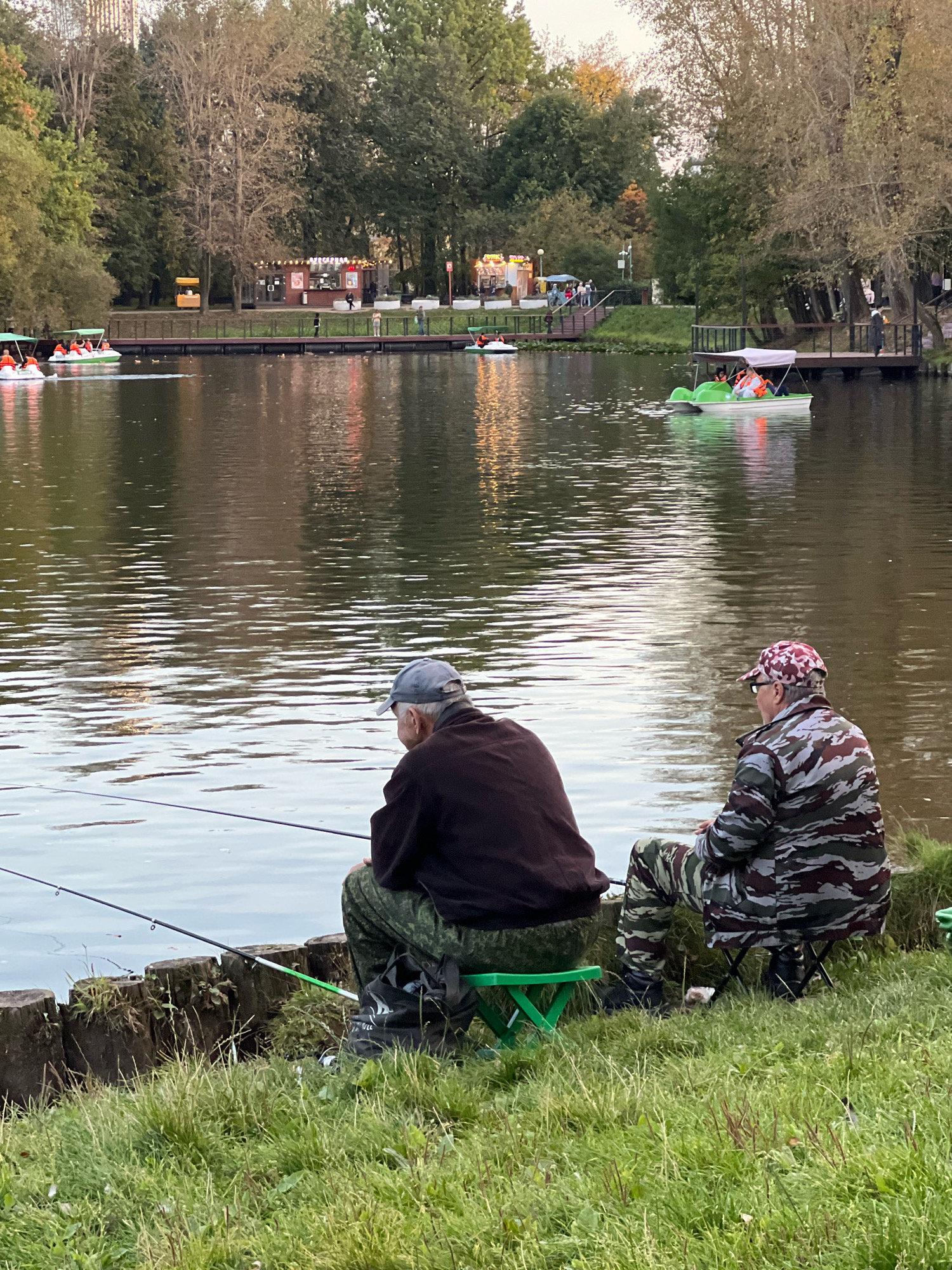 This is a full-shot, eye-level photograph taken outdoors in what appears to be a park setting in Moscow, Russia, likely during autumn. The scene depicts a tranquil body of water with reflections of trees and structures. In the foreground, two individuals are seated on collapsible stools, fishing. The person on the left is wearing a dark jacket, camouflage pants, and a light-colored cap. The person on the right is wearing a camouflage jacket, camouflage pants, and a camouflage cap with glasses. Both have fishing rods cast into the water.
In the mid-ground, several paddle boats are visible on the lake. Some are occupied by people wearing orange life vests. A wooden walkway or pier runs along the edge of the water, with small kiosks or food stands visible on the embankment behind it. People are seen walking along the pier and the embankment.
The background consists of a dense line of trees, some of which are displaying autumn colors. The sky is overcast, and the overall lighting suggests it is either early morning or late afternoon. The environment appears to be a public recreational area, possibly a park with a pond or lake, a common feature in many of Moscow's larger parks like Gorky Park or Sokolniki Park. The presence of paddle boats and fishing activities indicates a leisure-oriented location.