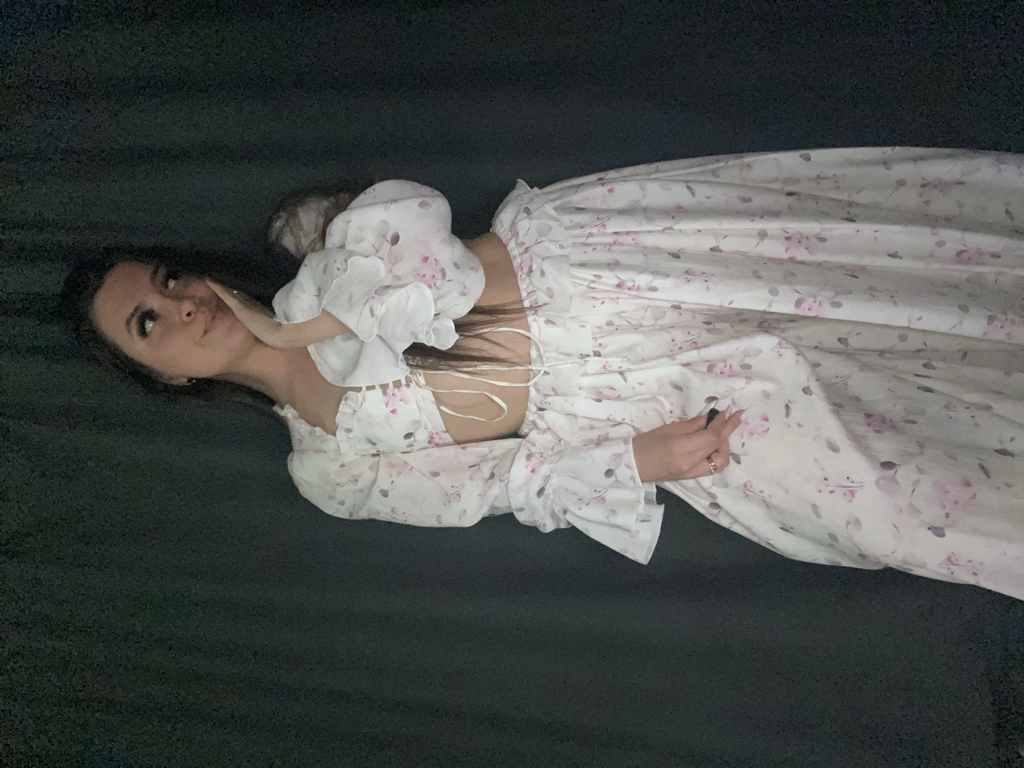 A young woman lies on a dark surface. She wears a floral print dress with a white background and light pink flowers. The dress has a ruffled neckline and puffed sleeves. Her dark hair is loose around her face. She is looking upwards and to the left with her hand near her cheek. Her left hand is near her waist. The location is not discernable and cannot be located within Chaikovsky, Russia.
