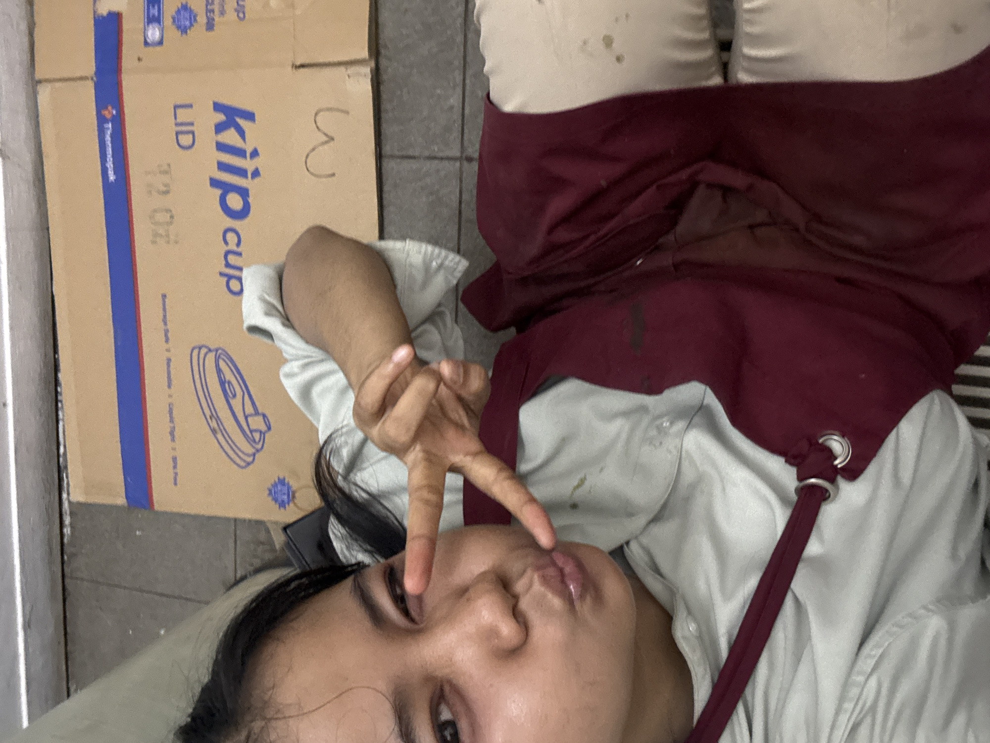 A person is lying on a tiled floor next to a cardboard box labeled "Klipp Cup Lid 12 oz." The box has the Thermopak logo and a large number "3" written on it. The person is wearing light-colored pants and a burgundy apron over a light gray shirt. The apron is stained. They are making a peace sign with their hand near their face, which has a pouting expression. The setting appears to be an indoor location in a city. The location in the city is not determinable from the image. It is unknown if the location is in North Jakarta, Indonesia.