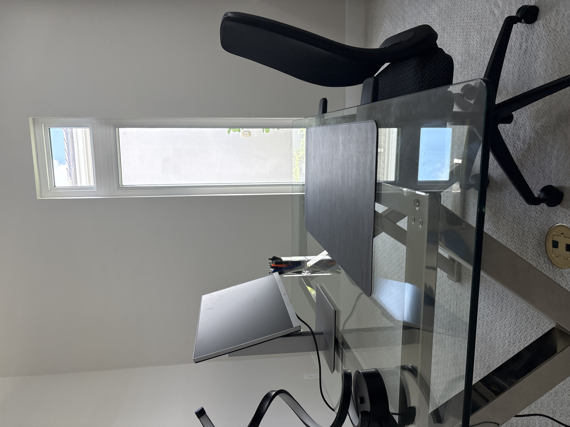 An interior, eye-level, medium shot captures a modern home office setup. The scene is dominated by a sleek glass-top desk with a chrome frame, reflecting the light and surrounding environment. Positioned on the desk is a silver computer monitor, a black desk lamp with a sculptural design, and a clear plastic container holding an assortment of pens and pencils. A black desk mat sits in front of the monitor. To the right of the desk, a black ergonomic office chair with a mesh back is visible. The backdrop consists of a white wall and a tall, narrow window with a white frame, offering a glimpse of the sky and the top of a building or roof. The floor is covered in light-colored, textured carpeting. The overall impression is one of a clean, contemporary workspace. The location is Bradenton, United States.