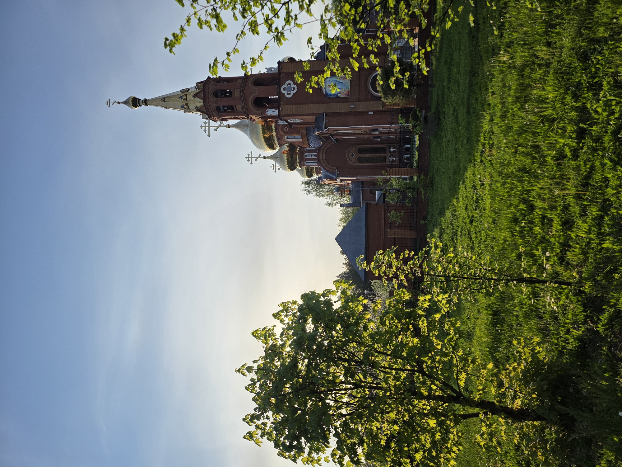 The image shows a brick church with golden domes and a pointed tower, likely located in the city of Chaikovsky, Russia. The church is surrounded by green grass and trees with green leaves. The sky is blue with some haze near the horizon. The building appears to be an Orthodox church with crosses on top of its domes and tower.