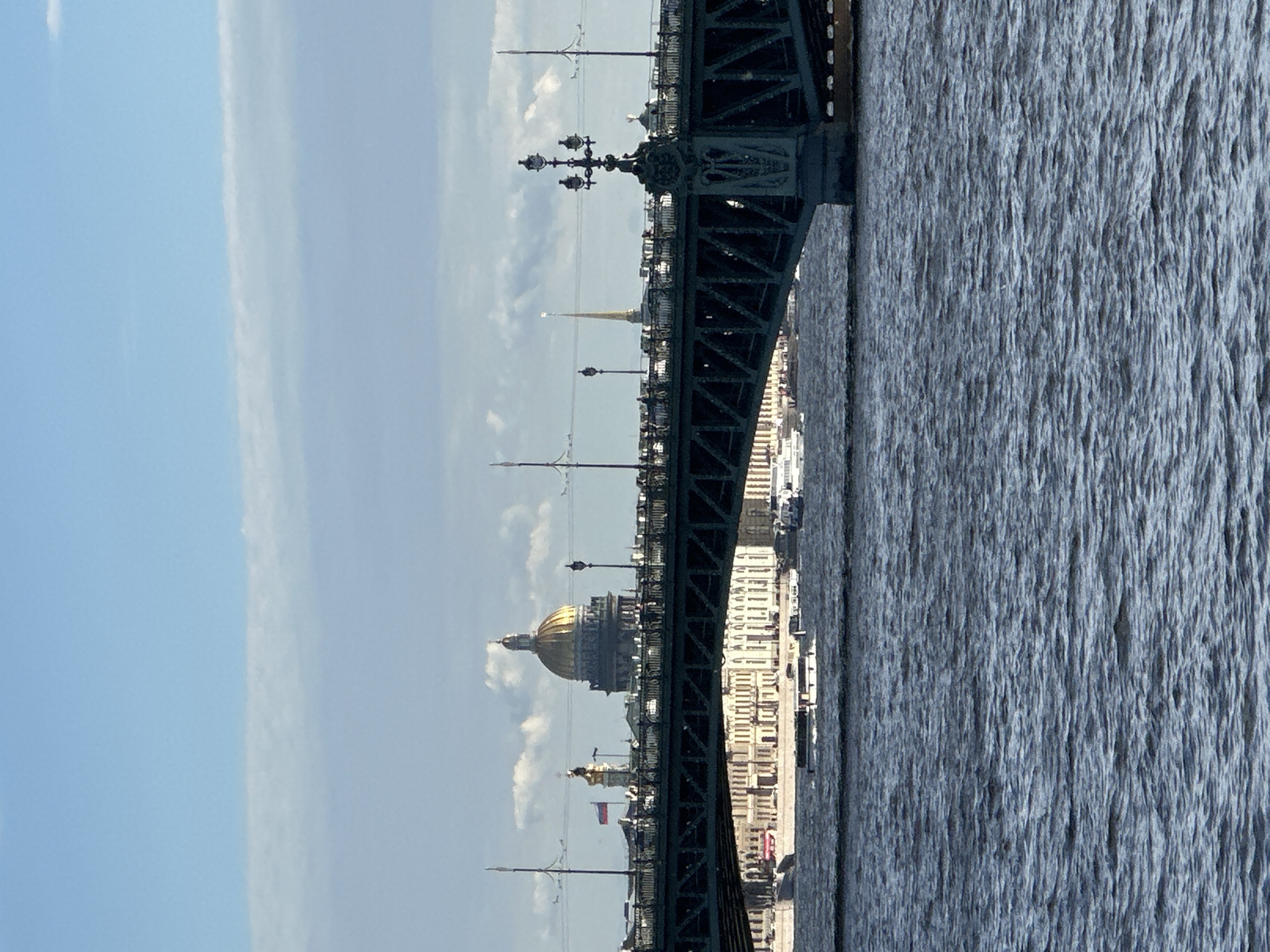 The image shows a view from a bridge overlooking a city skyline and a body of water. The perspective is vertical, suggesting the photo was taken while standing on the bridge. The bridge itself is a prominent feature, with dark green metal latticework and decorative lampposts. In the background, a city skyline is visible, characterized by numerous buildings and a distinctive golden dome of a large church or cathedral. A slender, spire-like tower also stands out against the sky. The water in the foreground is choppy, indicating some movement, possibly from wind or boat traffic. The sky is a clear blue with some wispy white clouds. The overall lighting suggests it is daytime, likely late morning or early afternoon. The scene appears to be of Saint Petersburg, Russia, given the architectural style and the presence of what looks like St. Isaac's Cathedral with its golden dome. No people are clearly visible, and no text is discernible. The mood is serene and picturesque, capturing a classic urban waterfront vista.