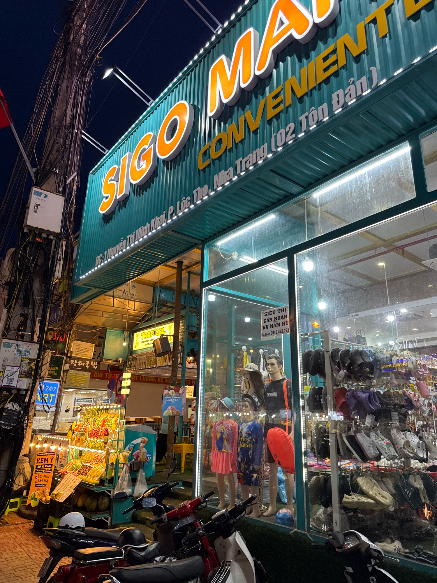 This is a nighttime exterior, eye-level, medium shot of a "Sigo Mart Convenient Store" in Nha Trang, Vietnam. The store's teal corrugated metal facade is illuminated by a string of white LED lights along its edge. Large, orange, three-dimensional letters spelling "SIGO MA" are prominent on the facade, with "CONVENIENT" in yellow letters beneath it. Below this, white text reads "202 Nguyễn Thị Minh Khai, P. Lộc Thọ, Nha Trang (02 Tôn Dân)". The store has a large glass storefront displaying mannequins in casual clothing, and various types of footwear are hung on display racks inside. To the left of the store, a busy street scene is visible. An illuminated fruit stand is set up on the sidewalk, with stacks of fresh produce. Several motorbikes are parked on the sidewalk in front of the store and fruit stand. Overhead, a dense network of electrical wires crisscrosses the dark sky, a common sight in Vietnamese cities. The overall atmosphere is bustling and vibrant, characteristic of a lively urban environment at night.