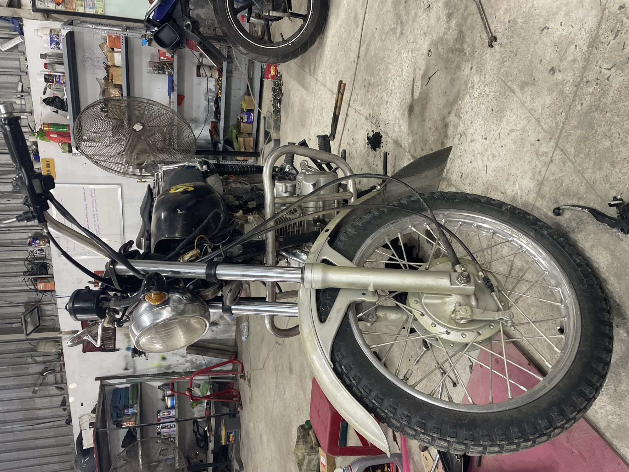 The image shows a motorcycle in a repair shop in Bac Nha Trang Ward, Vietnam. The motorcycle is partially disassembled, with its front wheel prominently displayed. Other motorcycles and parts are visible in the background, along with shelves holding tools and supplies. A fan stands behind the motorcycle's engine. The floor is concrete and shows signs of use.