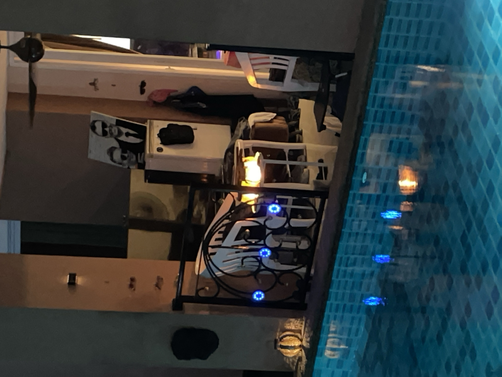 The image depicts a nighttime scene at a poolside in Pattaya City, Thailand. In the foreground, a swimming pool with blue tiled walls is visible, with several blue underwater lights creating illuminated reflections. Beyond the pool, a black metal railing with intricate scrollwork separates the pool area from a patio or balcony. Attached to the railing are several blue circular lights.
On the patio, a round, decorative light fixture with a striped pattern sits on a surface. In the background, there is a glimpse of indoor space with a tall white cabinet. A framed picture of two people wearing sunglasses is displayed on top of the cabinet. Various objects, including chairs, a small table with a lit lantern or heater, and hanging clothing, are scattered around the patio area. The overall ambiance suggests a leisure or entertainment setting.
