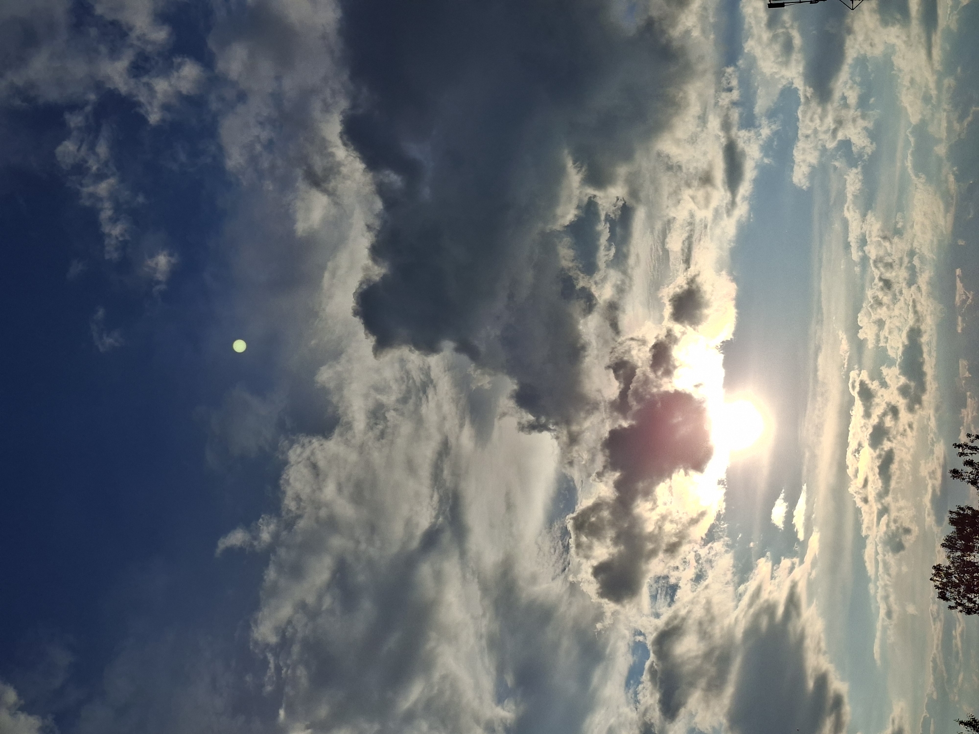 The image depicts a daytime sky filled with clouds. A bright sun is visible, partially obscured by clouds. The sky ranges from dark blue to lighter hues. There is also a light green orb visible in the dark blue section of the sky. Trees are present on the right side. The image appears to be taken looking upwards. The location is Unknown, Unknown.