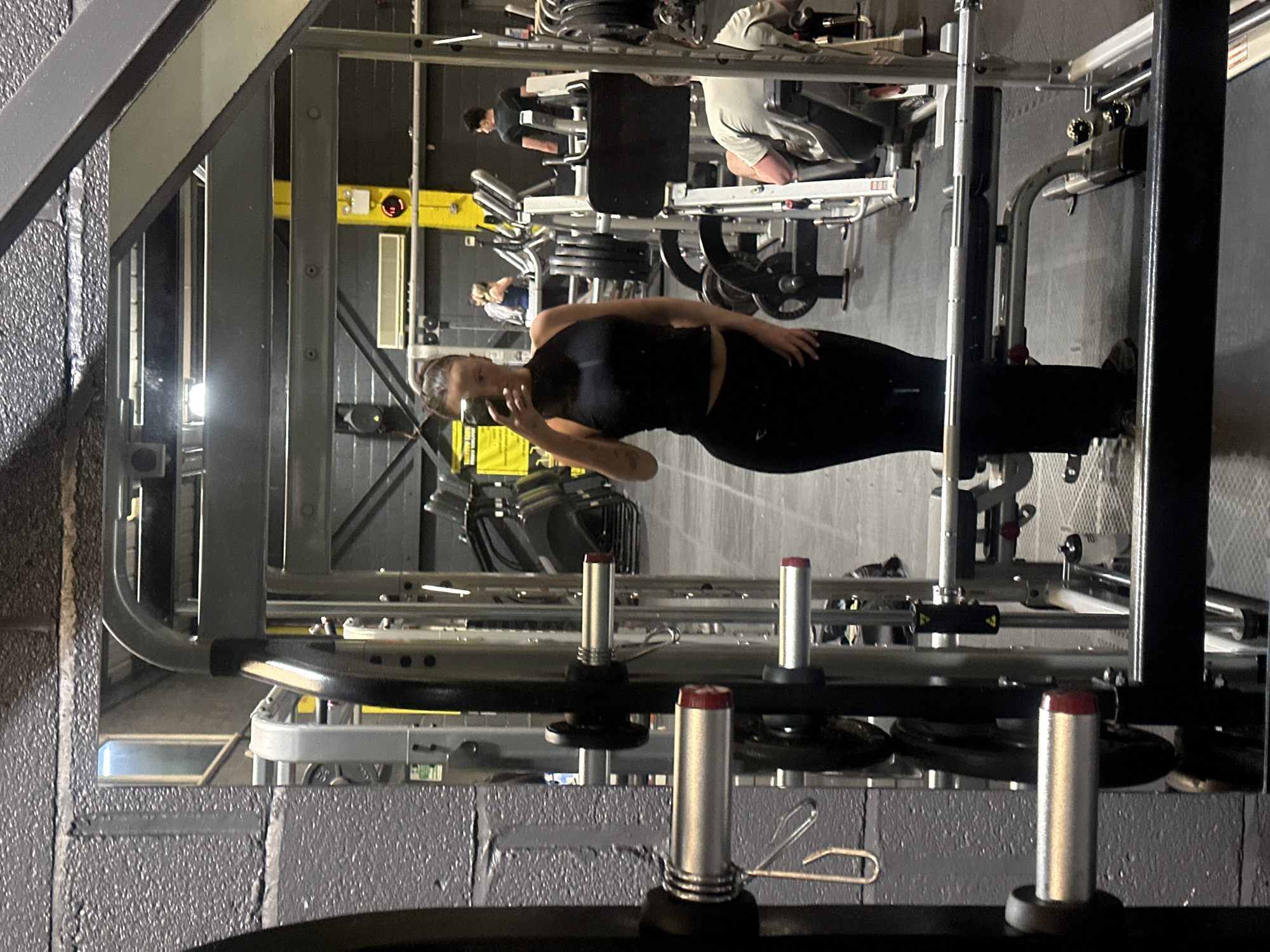 The image depicts the interior of a gym, specifically Invictus Gym located in Tonbridge, Kent, within the Borough of Tonbridge and Malling, United Kingdom. The scene is observed through a large reflective surface, likely a mirror, with a dark grey textured wall and a silver-framed squat rack or Smith machine in the immediate foreground, creating the frame for the reflection.
In the reflected view, a female individual, presumed to be the photographer, stands centrally, capturing a mirror selfie with a smartphone held in both hands. She wears a black sleeveless crop top and matching black fitted trousers, with her dark hair pulled back. A tattoo is visible on her left upper arm.
The gym environment exhibits an industrial aesthetic with dark grey walls and a dark floor. Overhead, light fixtures and exposed metal beams are visible. In the mid-ground, a prominent yellow vertical structural beam features a red digital display indicating "12". To the left of this beam, a yellow sign is partially obscured, and a white horizontal ventilation unit is mounted on the wall.
Several other individuals are present in the background. To the right, a male is seated on a leg press machine, facing away from the camera, wearing a light-colored t-shirt. Another male stands further back and to the right, wearing a dark t-shirt. In the distant left background, a female is visible on a cardio machine, wearing a blue top. Various gym equipment, including weight machines, treadmills, and stacked weight plates, populate the area. A black "Elite" branded water bottle rests near the leg press machine.