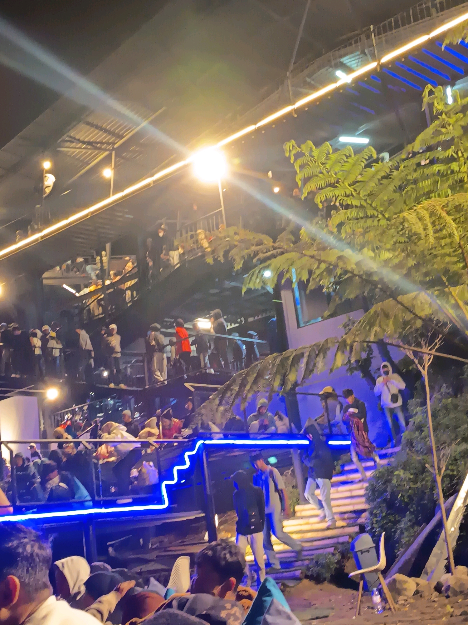 The image depicts a nighttime scene at a multi-level outdoor venue, likely a cafe or restaurant, in Keditan, Indonesia. The venue is illuminated by artificial lighting, including yellow string lights along the railings and stairs, and bright blue neon lights accentuating the edges of the upper levels. A large, leafy green plant, possibly a fern, dominates the right side of the frame, its fronds reaching across the scene.
Numerous people are present, distributed across the various levels of the venue. Many appear to be standing or walking along stairs and walkways. Some individuals are dressed in casual attire, including hoodies, jackets, and head coverings. Several people are gathered on a central staircase, while others are visible on balconies and walkways above and below. The atmosphere appears to be lively, with a crowd of patrons enjoying the evening. The ground level is rough, with some rocks and dirt visible. A blue chair is positioned on the right side of the ground level, with a plastic bottle resting at its base. The overall impression is of a popular social gathering spot at night.
