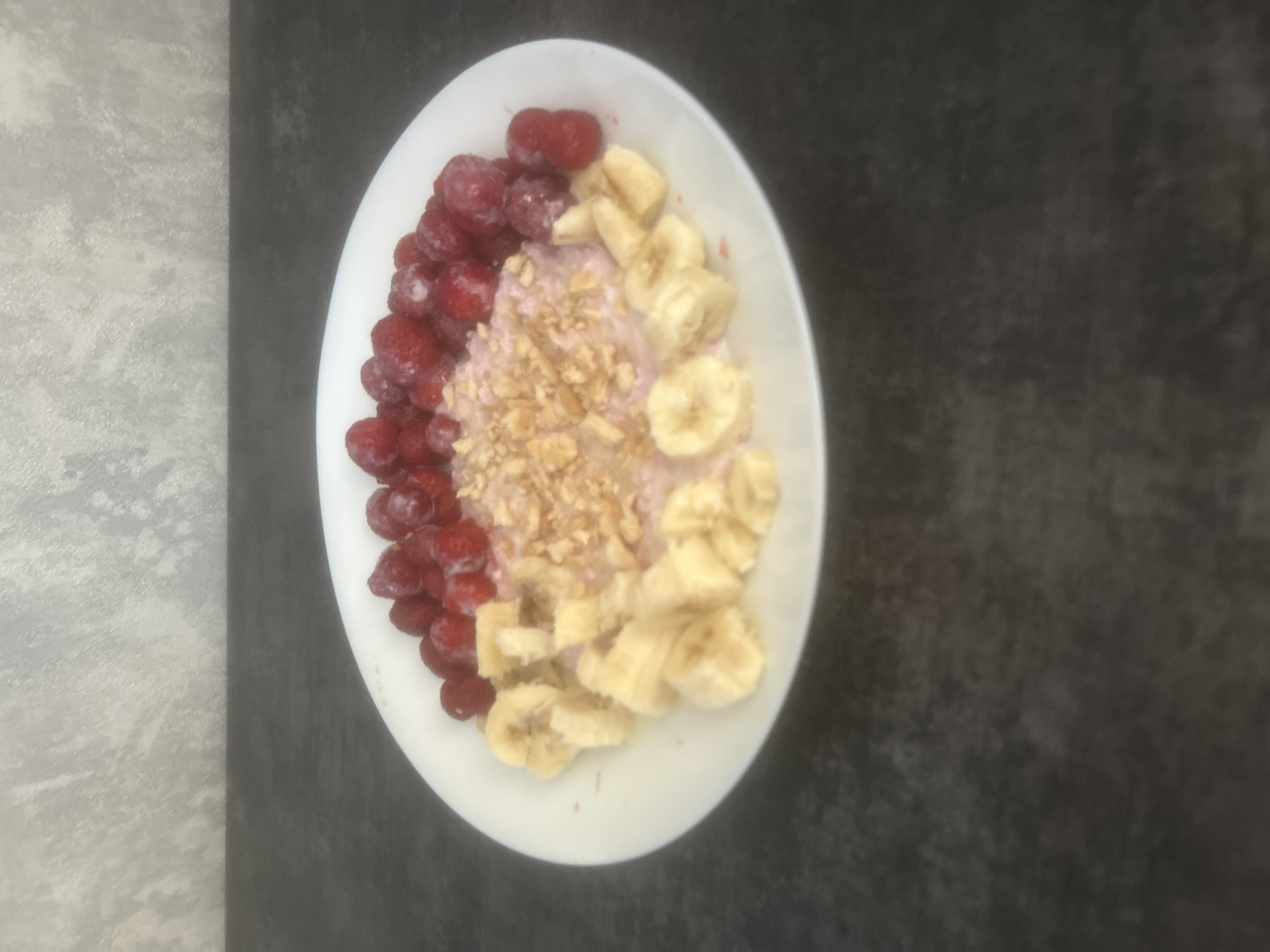 A white oval bowl of oatmeal is presented from a top-down perspective, rotated to the left. The oatmeal is topped with frozen raspberries on the left, sliced bananas on the right, and sprinkled with crushed nuts in the center. The bowl is placed on a dark, textured surface that appears to be a countertop. To the left of the bowl, there is a lighter, mottled grey surface, possibly another section of the countertop or a placemat. There are no people, text, or other distinguishing features in the image. The lighting suggests an indoor setting, potentially during the daytime. The overall impression is that of a healthy breakfast.