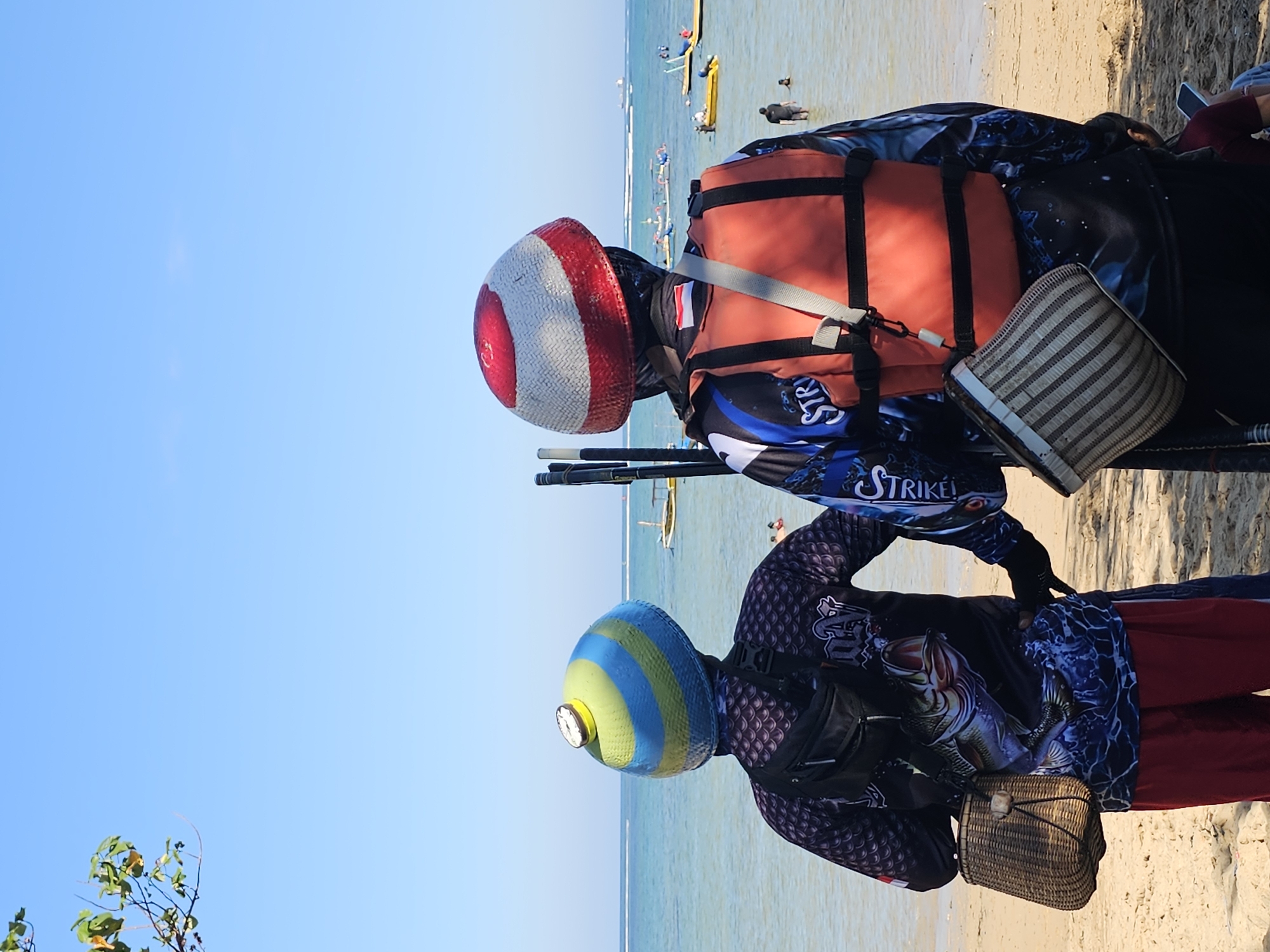 Two individuals stand facing away from the camera on a sandy beach, with the ocean and several small boats in the background. The person on the left wears a black long-sleeved shirt with a large fish graphic, red pants, and a blue, green, and yellow conical hat. They have a black fanny pack and a woven basket attached to their side. The person on the right wears a black long-sleeved shirt with blue and white graphics, an orange life vest, and a red and white striped conical hat. They also have a woven basket attached to their side. The sky is clear and blue, and the water appears calm. The scene suggests a sunny day at a coastal location, potentially a tourist area within Denpasar, Indonesia, given the presence of boats and people in the water.