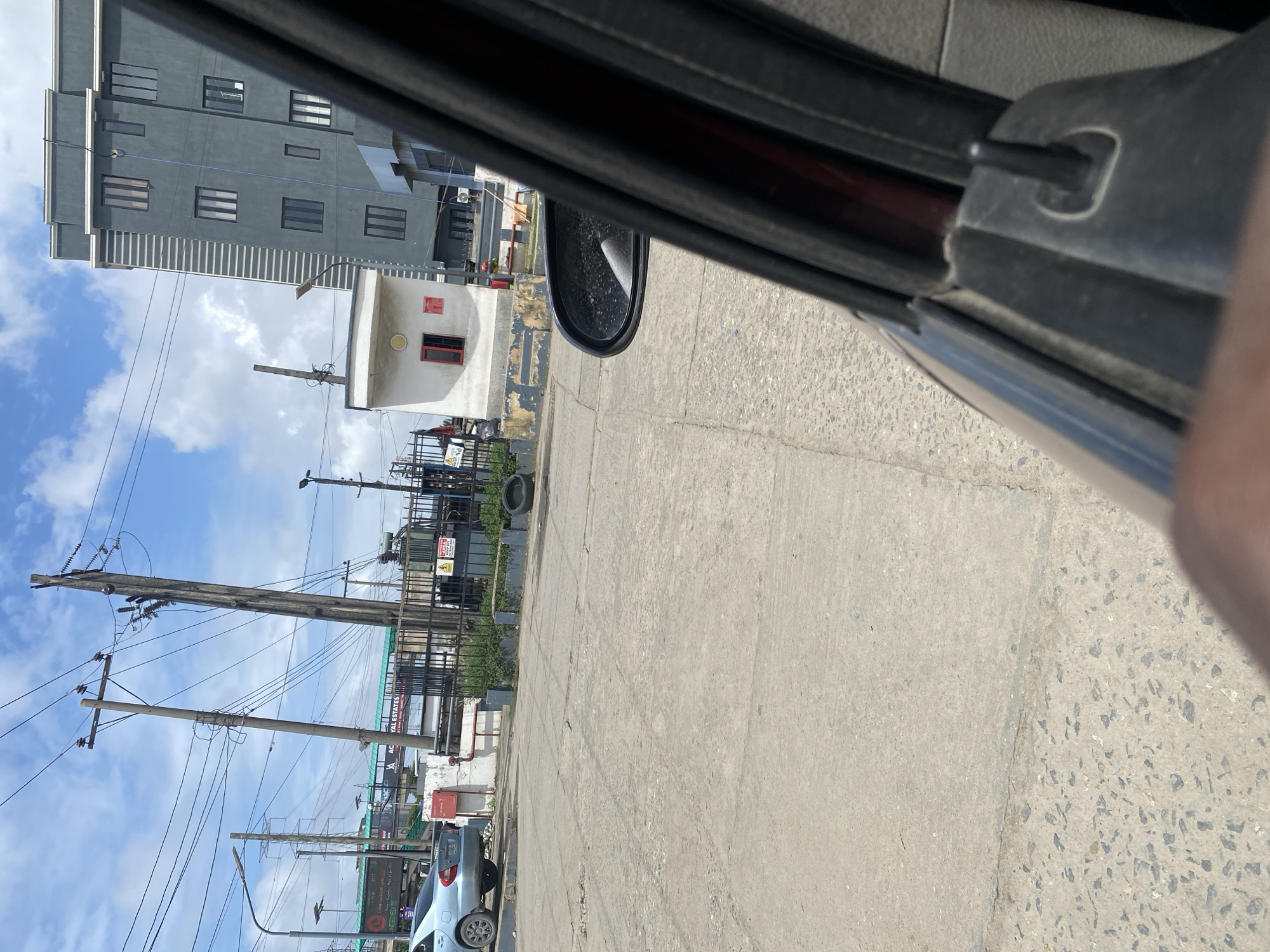The image depicts a view from inside a vehicle looking out at a street scene in Ajah, Nigeria. In the foreground, the interior of the car, including a portion of the door panel and a side mirror, is visible. The exterior of the car shows a light blue sedan parked on the left side of the frame.
The street is paved with concrete and appears to be in moderate condition with some cracks and wear. In the middle ground, several large, wooden utility poles with extensive electrical wiring are prominent. Adjacent to these poles is a fenced-off area containing a large electrical transformer and a white building with a red-framed window. Another, taller modern building with multiple windows stands in the background to the right.
Further back, a pedestrian bridge can be seen spanning over the road, with advertisements displayed on its sides. The sky is partly cloudy, suggesting daytime conditions. The overall environment appears to be a mix of urban infrastructure and some commercial or residential development.