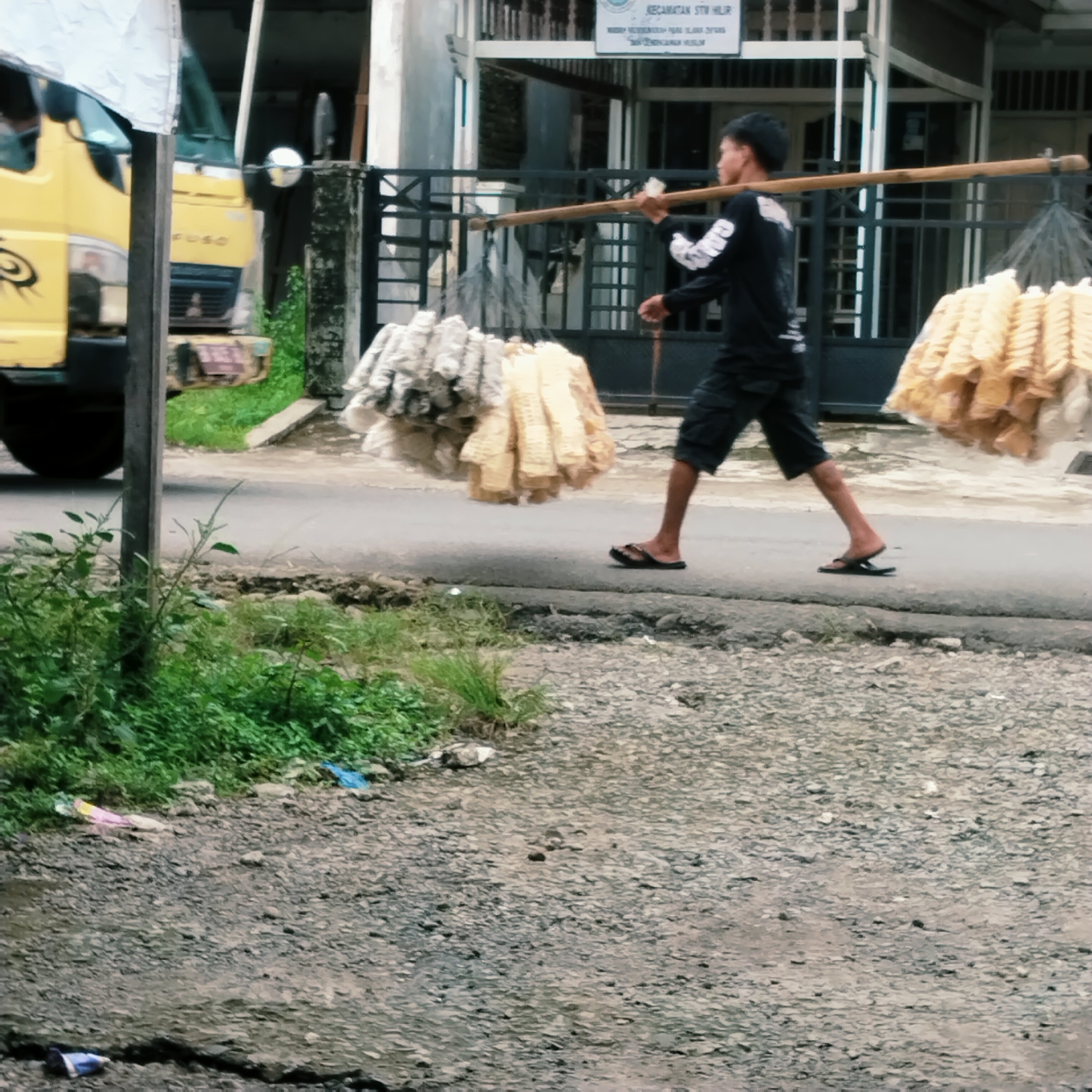 In the city of Talun Kenas, Indonesia, a man in a black t-shirt and shorts walks on a paved road, carrying a long wooden pole balanced on his shoulders. Attached to the ends of the pole are bundles of light-colored, possibly fried, food items wrapped in plastic. To the left of the man, a yellow truck is partially visible, suggesting an urban or semi-urban setting. The roadside is lined with green vegetation and some scattered debris, including pieces of plastic. The ground is a mixture of gravel and cracked pavement. The scene appears to be during daylight hours.