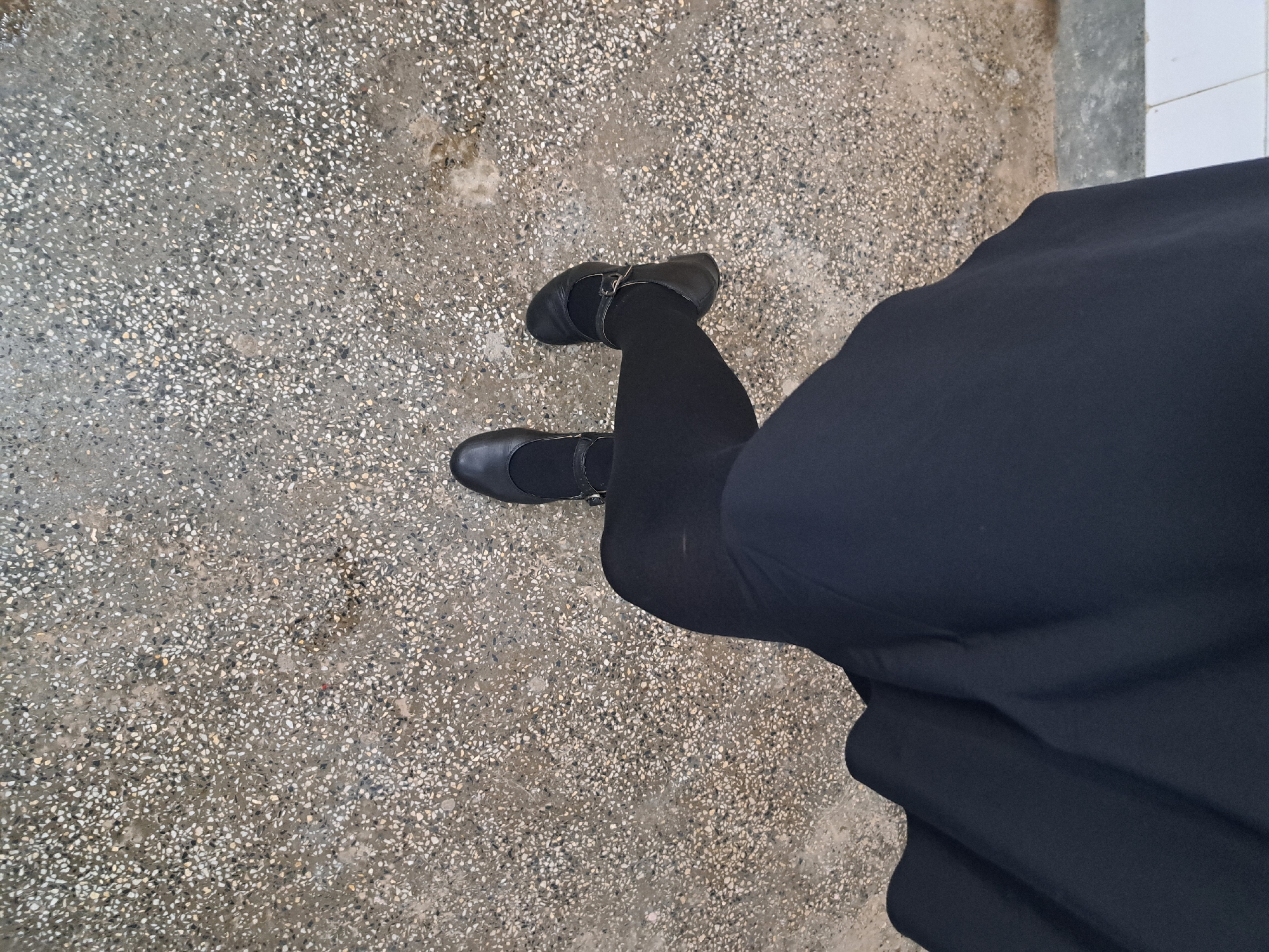 The image, taken in Lokorsko, Bulgaria, presents a high-angle, overhead view of a person's lower body, standing or walking on a speckled ground surface. The individual is wearing black opaque tights or leggings, black flat shoes with a buckle strap across the instep (Mary Jane style), and a long black skirt or dress that extends downwards, covering a significant portion of their legs. The fabric of the garment appears smooth.
The ground beneath the person is a grey, textured surface, likely terrazzo or speckled concrete, composed of numerous small white, grey, and black aggregate chips. Some areas of the surface appear slightly damp or show variations in tone, possibly from moisture or wear. In the upper right section of the original image, a white tiled edge is partially visible, indicating a boundary with an adjacent structure or different flooring material. The lighting is bright and even, suggesting either natural daylight or ambient indoor lighting, as no distinct shadows are cast. There are no discernible activities beyond standing or movement. No text is visible in the image, and no specific time of day, weather conditions, or emotions can be determined from the provided visual information.