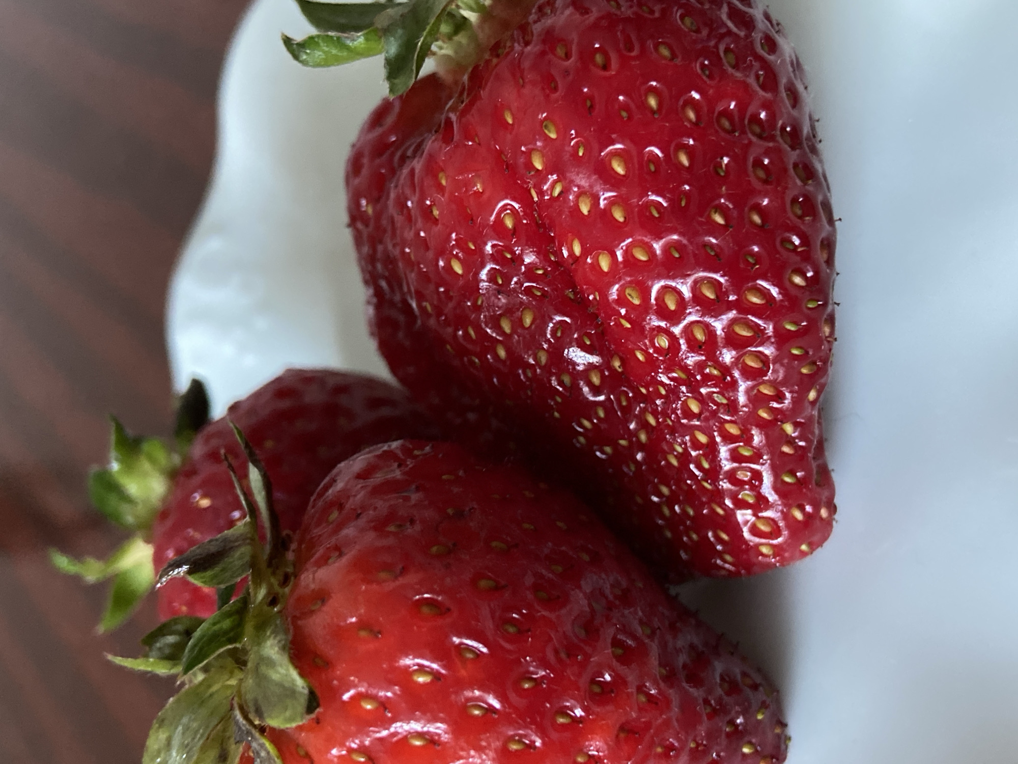 A close-up, high-angle shot features three vibrant red strawberries nestled in a white bowl, against a blurred dark brown background. The image is oriented vertically, with the strawberries occupying the majority of the frame.
The strawberries are plump and appear freshly washed, with a glossy, moist surface that reflects light. Their distinct texture is visible, showing numerous small, light yellow or greenish-yellow achenes (seeds) embedded across their bright red skin. Some of the strawberries still have their green calyxes and short stems attached, which are slightly faded or browning at the edges.
The white bowl has a gently curved or scalloped rim, visible at the top and left side of the original image, providing a soft contrast to the deep red of the fruit. The background is out of focus, revealing a warm, reddish-brown hue that suggests a wooden surface, likely a table. There is no visible text in the image.
