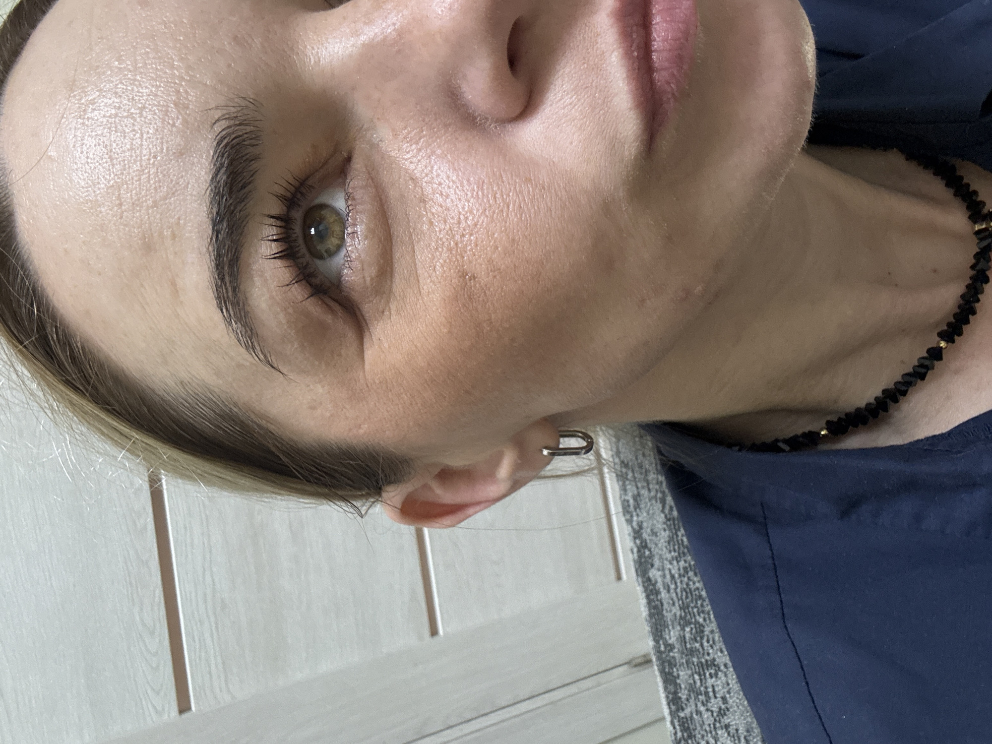 A close-up, rotated view of a fair-skinned individual, possibly a woman, with visible skin texture and fine lines. She has dark eyebrows, brown eyes with mascara, and light pink lips. She is wearing a necklace of small, irregular black beads with occasional gold-colored elements. A silver-colored hoop earring is visible in her left ear. She appears to be wearing a dark blue top. The background includes a light-colored paneled wall and a section of grey-patterned carpet. No location information in Unknown, Russia is discernible from the image.