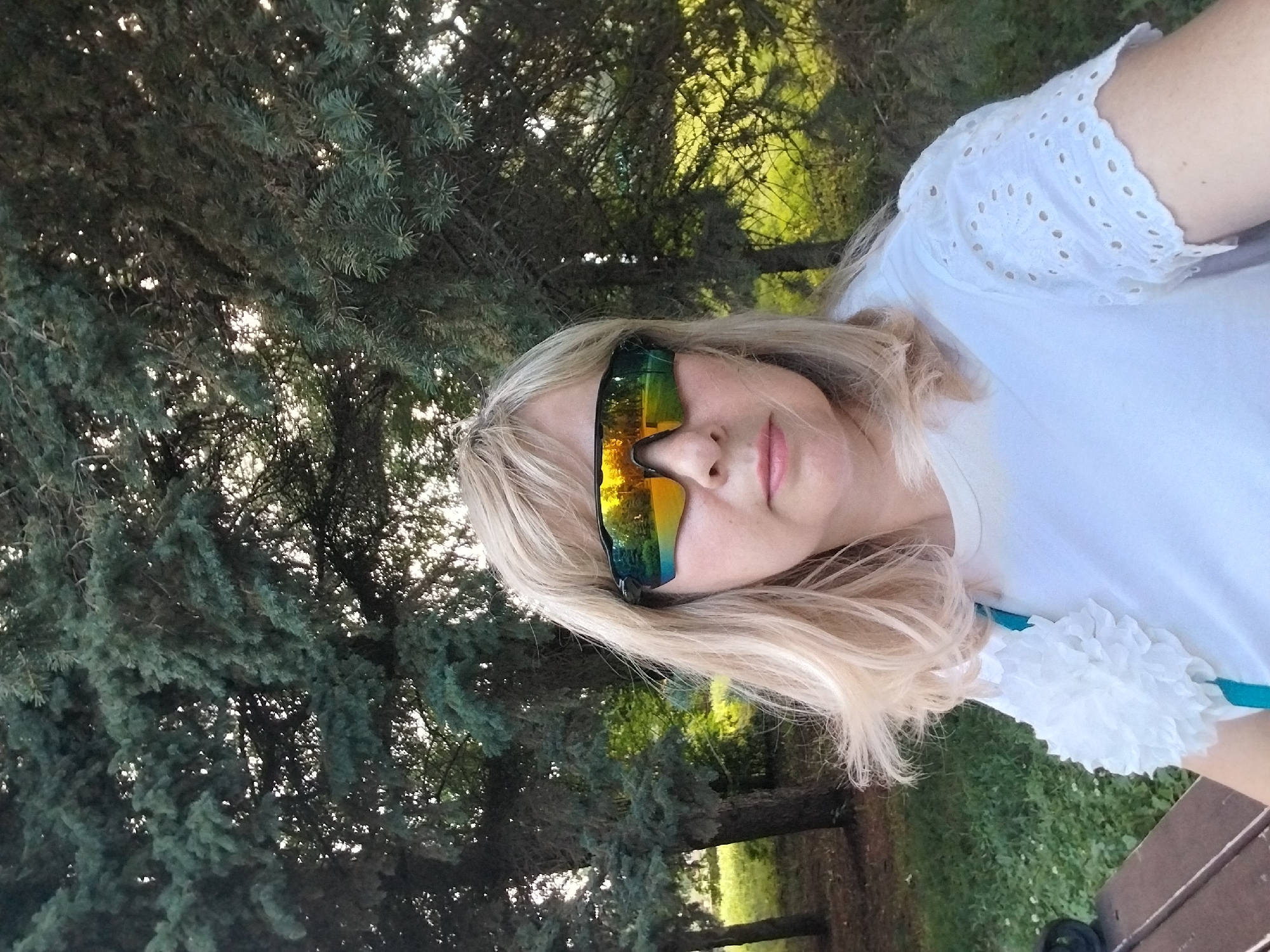A woman with blonde hair is taking a selfie outdoors. She is wearing sunglasses with mirrored lenses that reflect the surrounding trees and sky. She has on a white t-shirt with eyelet detailing on the sleeves. A white decorative flower is pinned to the left side of her shirt. She is partially seated on a wooden bench. The background consists of trees and green grass. The image appears to have been taken in the city of Orenburg, Russia.