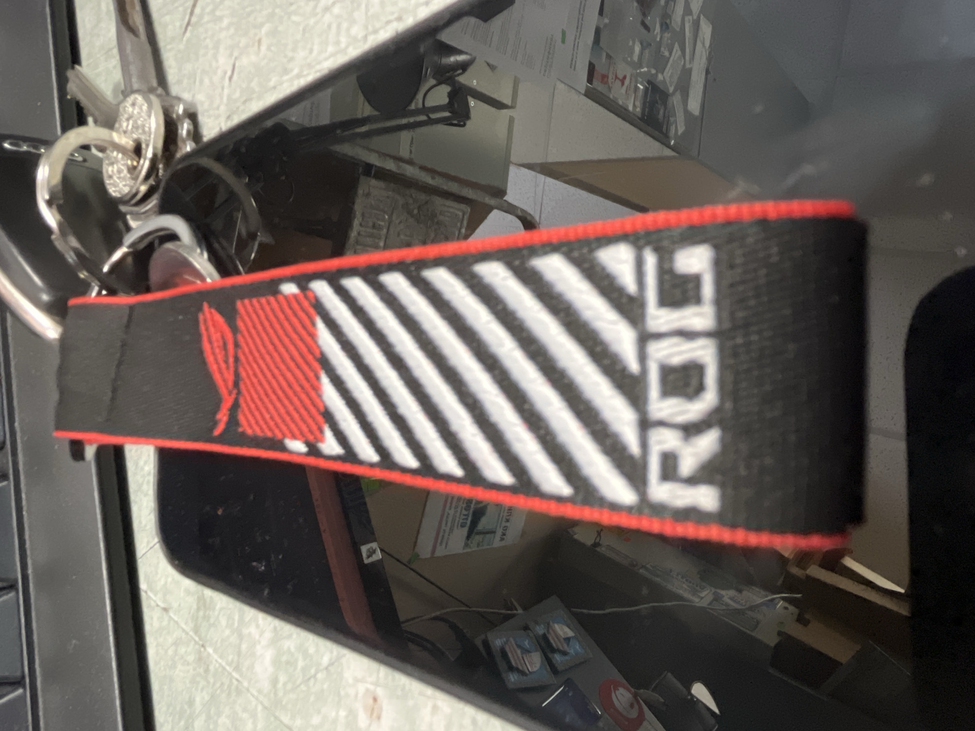A close-up shows a black key chain with the letters "ROG" in white, horizontal orientation, with alternating white and red stripes on a black background. The keychain has a red border. It is attached to a set of keys with a sparkly ring. The keychain and keys are lying on a black, reflective surface. Reflections of the surrounding area can be seen in the surface. Part of a computer keyboard is visible at the edge of the image.