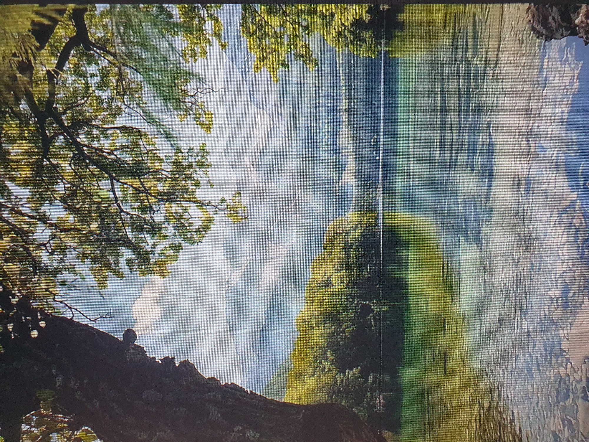 The image depicts a serene natural landscape. In the foreground, a clear, shallow body of water allows the viewer to see the rocky bottom beneath. The water's surface is calm, reflecting the surrounding environment. Beyond the water, lush green trees line the shore, forming a dense forest that stretches towards rolling hills. In the distance, a majestic mountain range rises, its peaks partially covered in snow and bathed in soft light. The sky above is a clear blue, with a few wispy clouds scattered across it. The scene evokes a sense of tranquility and natural beauty. This image is not of Tashkent, Uzbekistan.